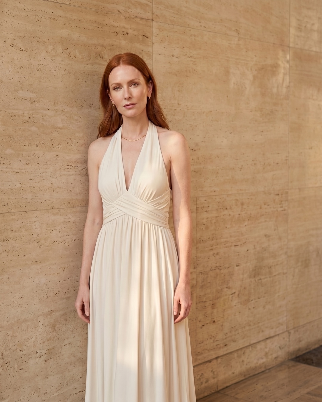 Hugo Caesar Tassi Aelia Grecian cross-front halter maxi dress in pale Avorio ivory, worn by a model beside fluted columns in a Roman travertine studio