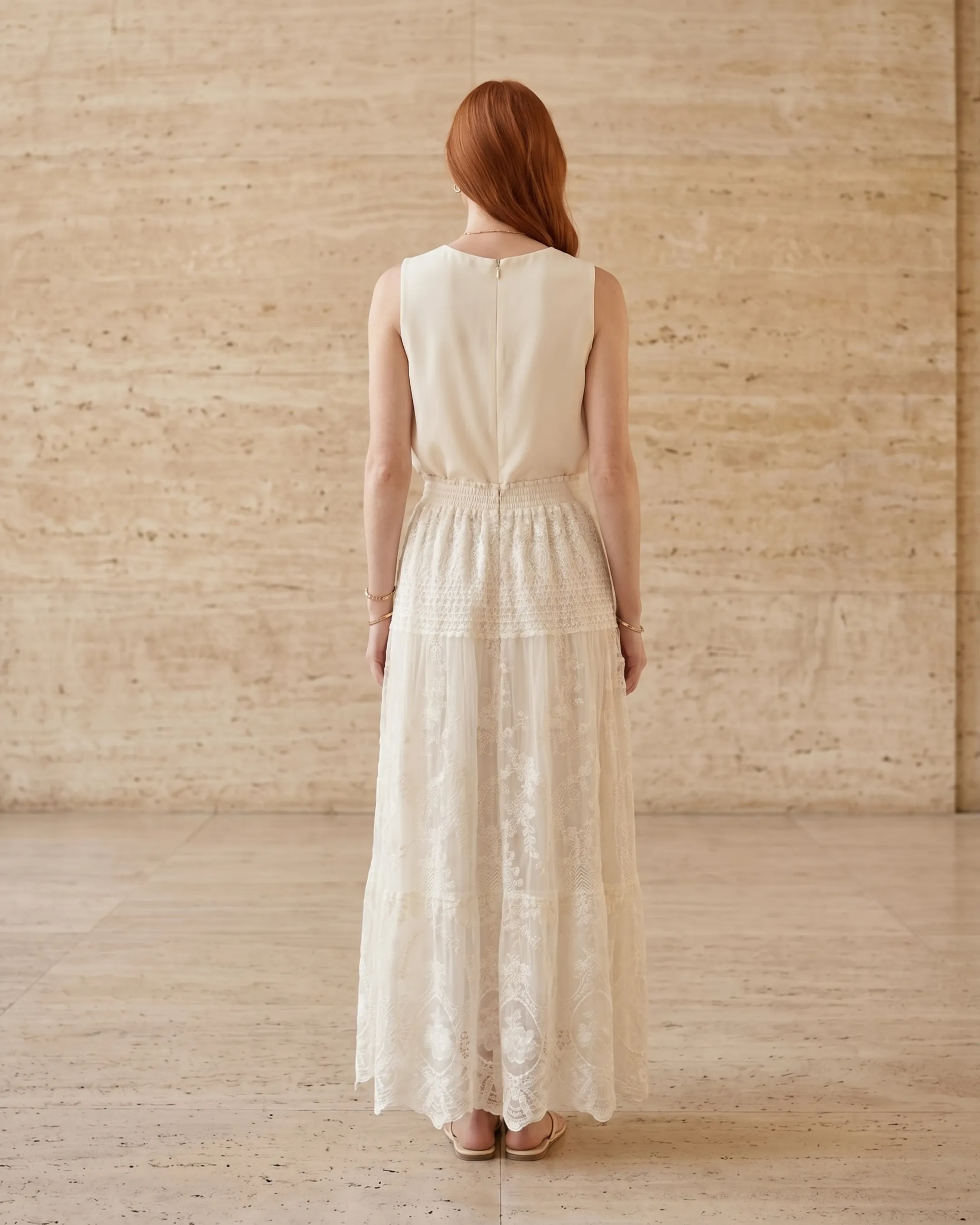 Full-length rear view of the Hugo Caesar Tassi Aemilia floral-embroidered lace A-line chiffon maxi dress in Avorio, seen from behind, showing the back of the dress, the sleeveless scoop back, shirred elastic waist and tiered scalloped-lace skirt sweeping the floor in a Roman travertine studio