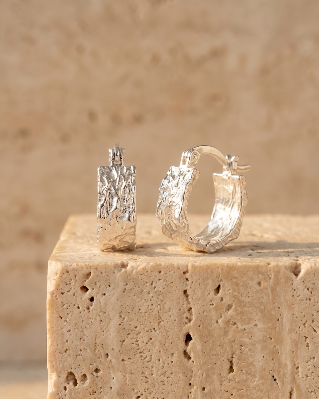 Hugo Caesar Tassi Argentea pair of wide-banded textured fine silver huggie hoop earrings resting on a warm travertine pedestal