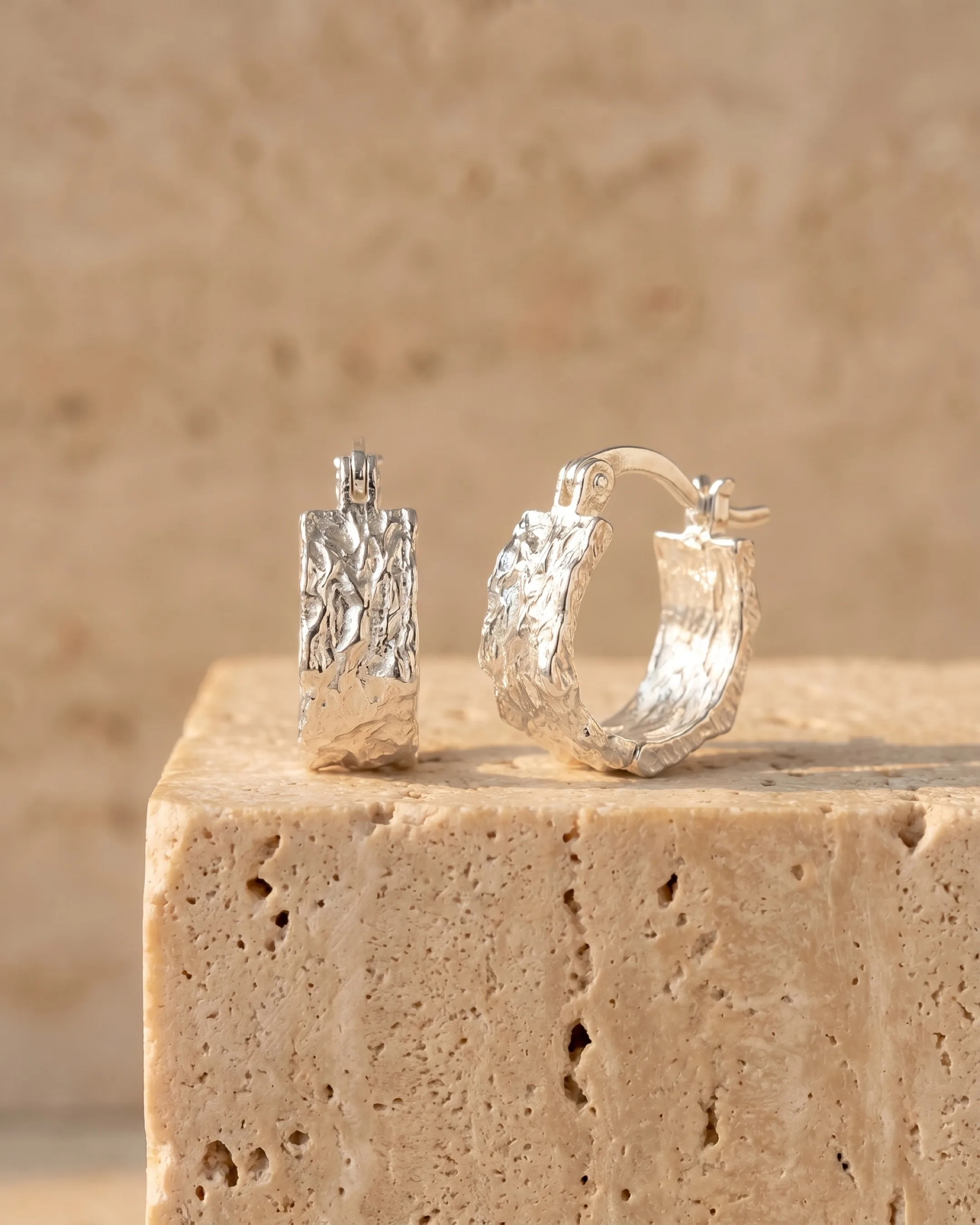 Hugo Caesar Tassi Argentea pair of wide-banded textured fine silver huggie hoop earrings resting on a warm travertine pedestal