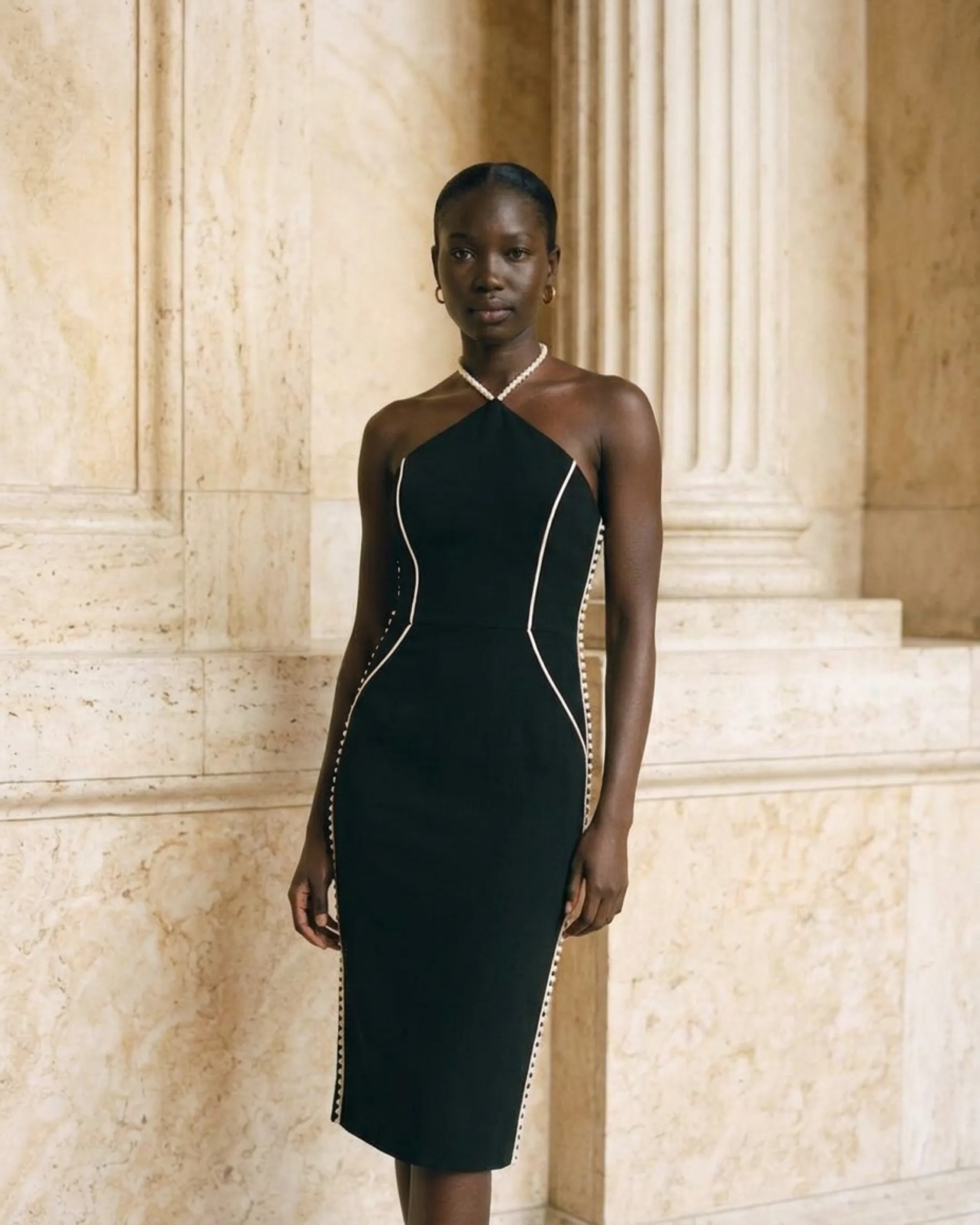 Hugo Caesar Tassi Caecilia editorial wide of the black sheath cocktail dress beside a fluted travertine column in golden light