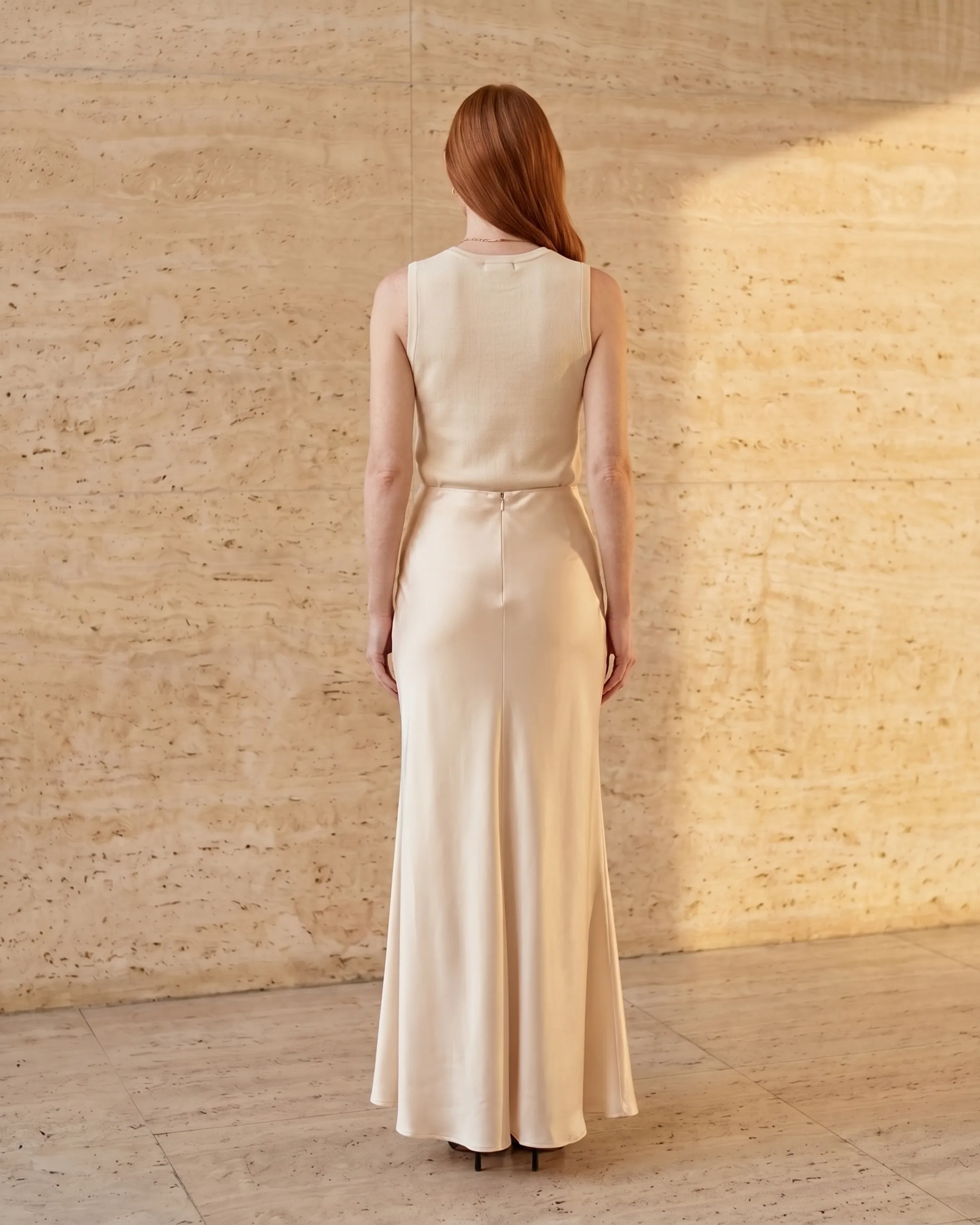 Full-length rear view of the Hugo Caesar Tassi Calpurnia fishtail satin evening gown in Avorio champagne, seen from behind, showing the back of the dress, the concealed back zip, high waist and bias-cut liquid-satin column flaring into the fluted mermaid fishtail in a Roman travertine studio