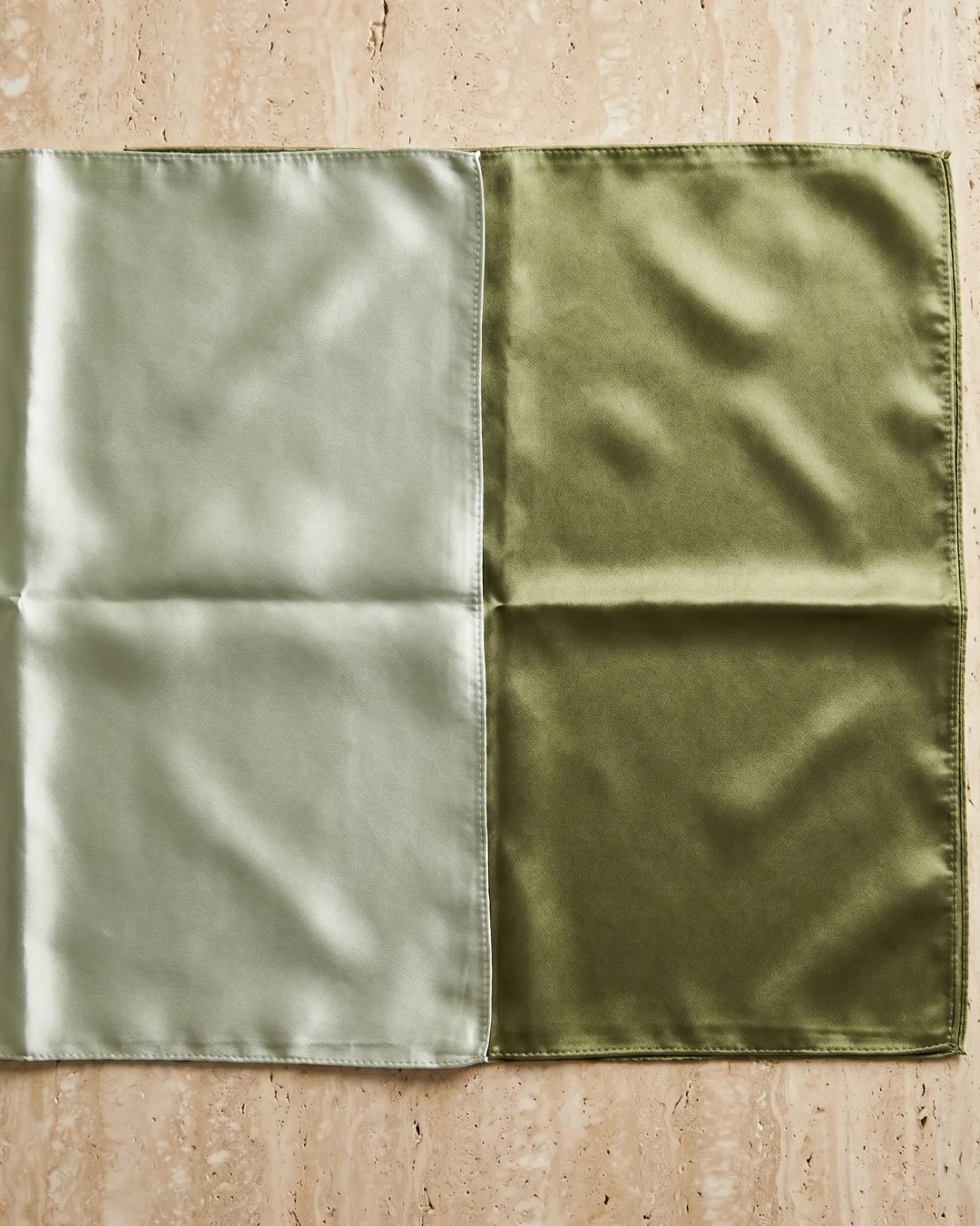 Hugo Caesar Tassi Carina glossy silk-look square scarf in Verde Antico laid open flat on a warm travertine-marble surface, showing the full two-tone glossy sheen edge to edge