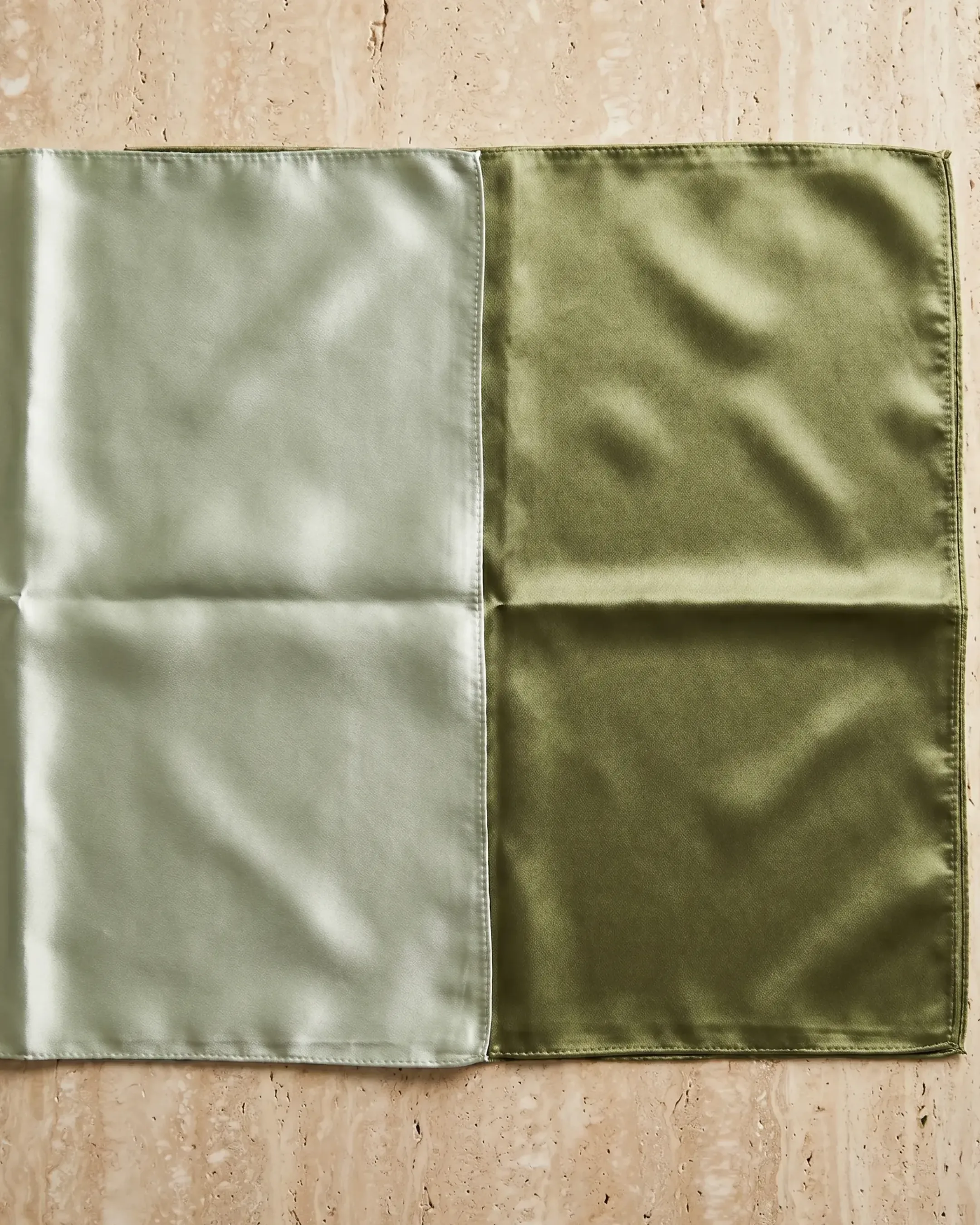 Hugo Caesar Tassi Carina glossy silk-look square scarf in Verde Antico laid open flat on a warm travertine-marble surface, showing the full two-tone glossy sheen edge to edge
