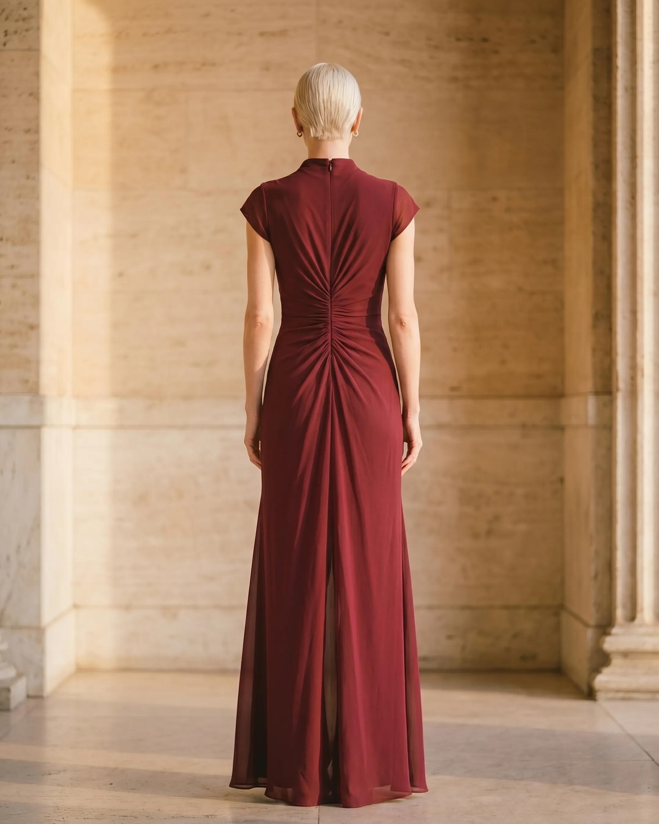 Full-length rear view of the Hugo Caesar Tassi Claudia pleated slit maxi evening gown in Porpora wine-red chiffon, seen from behind, showing the back of the dress, the high draped mock neck, ruched waist and floor-skimming column skirt in a Roman travertine studio