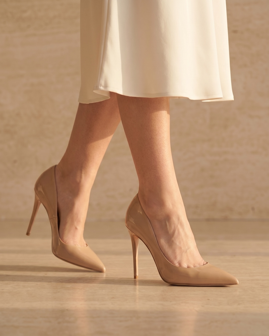 Hugo Caesar Tassi Crispina nude patent pointed pump worn on the foot below an ivory hemline against a fluted travertine wall