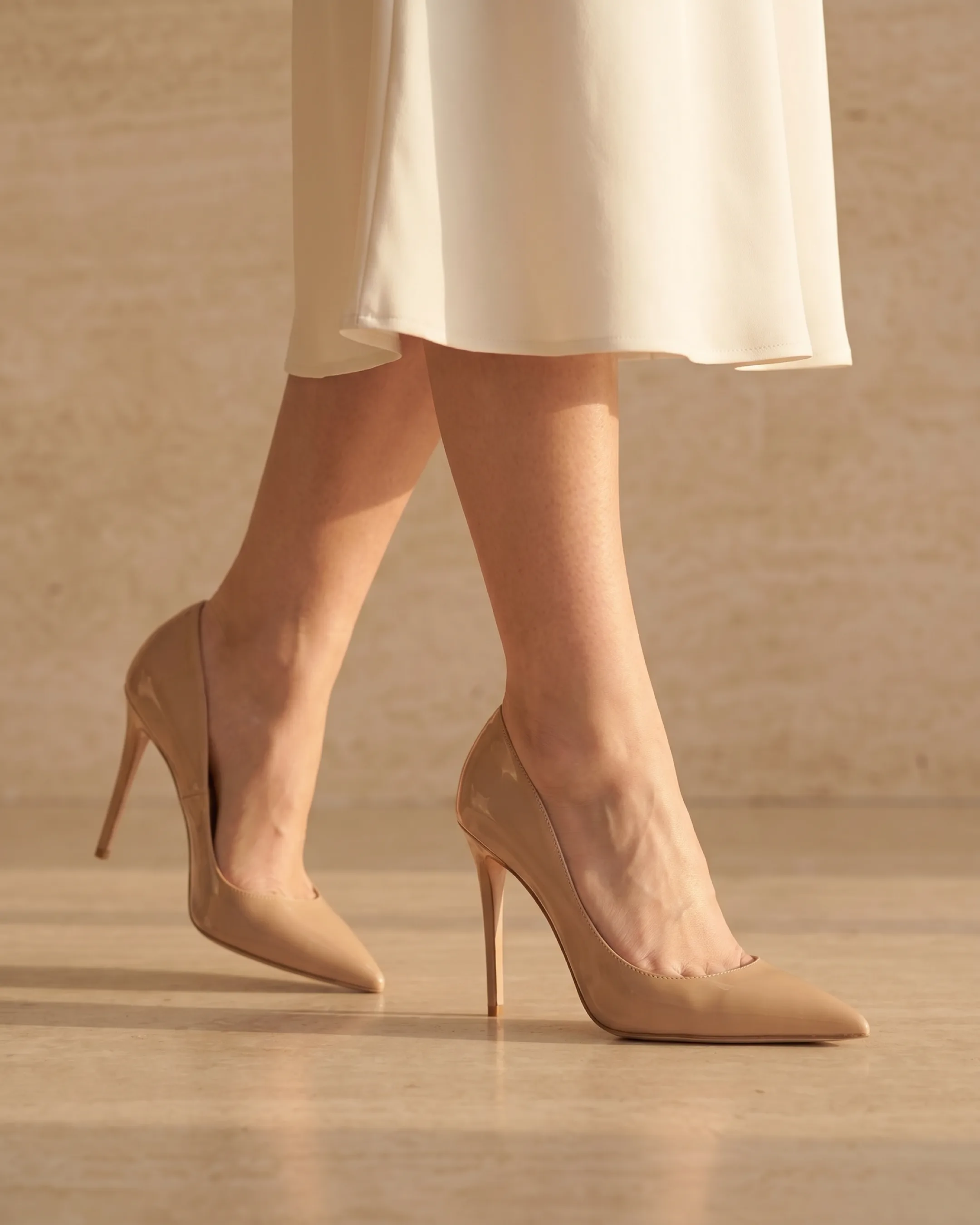 Hugo Caesar Tassi Crispina nude patent pointed pump worn on the foot below an ivory hemline against a fluted travertine wall