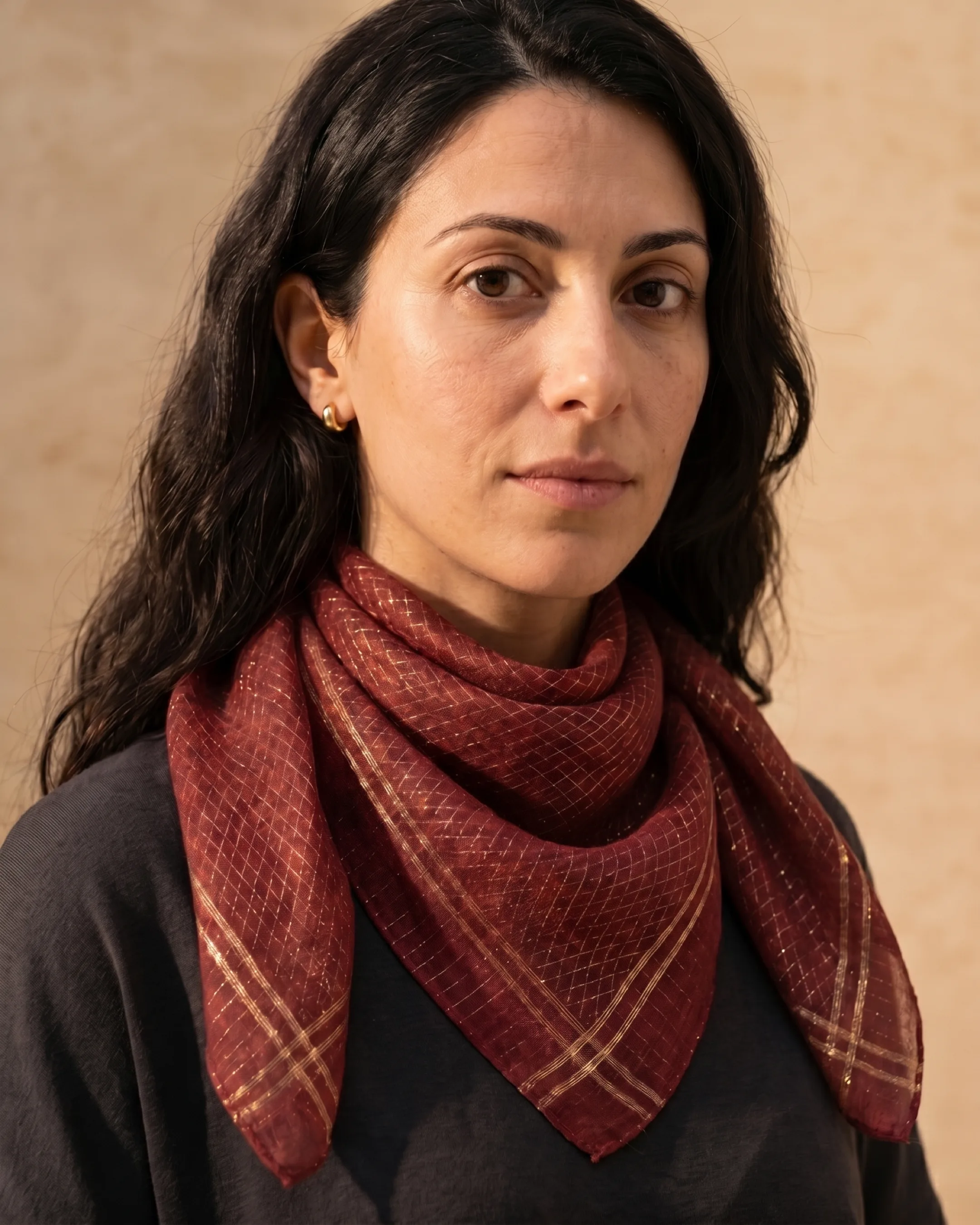 Hugo Caesar Tassi Cumae wine silk-wool scarf folded into a triangle at the throat of the model against a warm travertine wall, gold rhombus weave visible