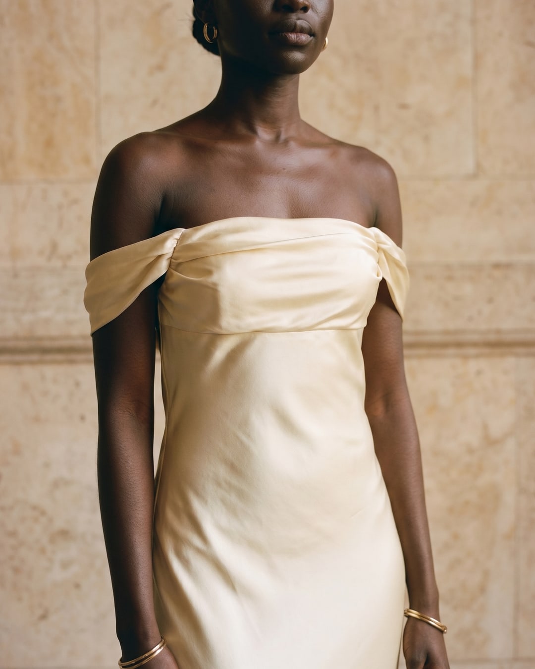 Off-the-shoulder draped satin bodice detail of the Hugo Caesar Tassi Flavia gown in champagne Avorio, lit by soft daylight against travertine marble
