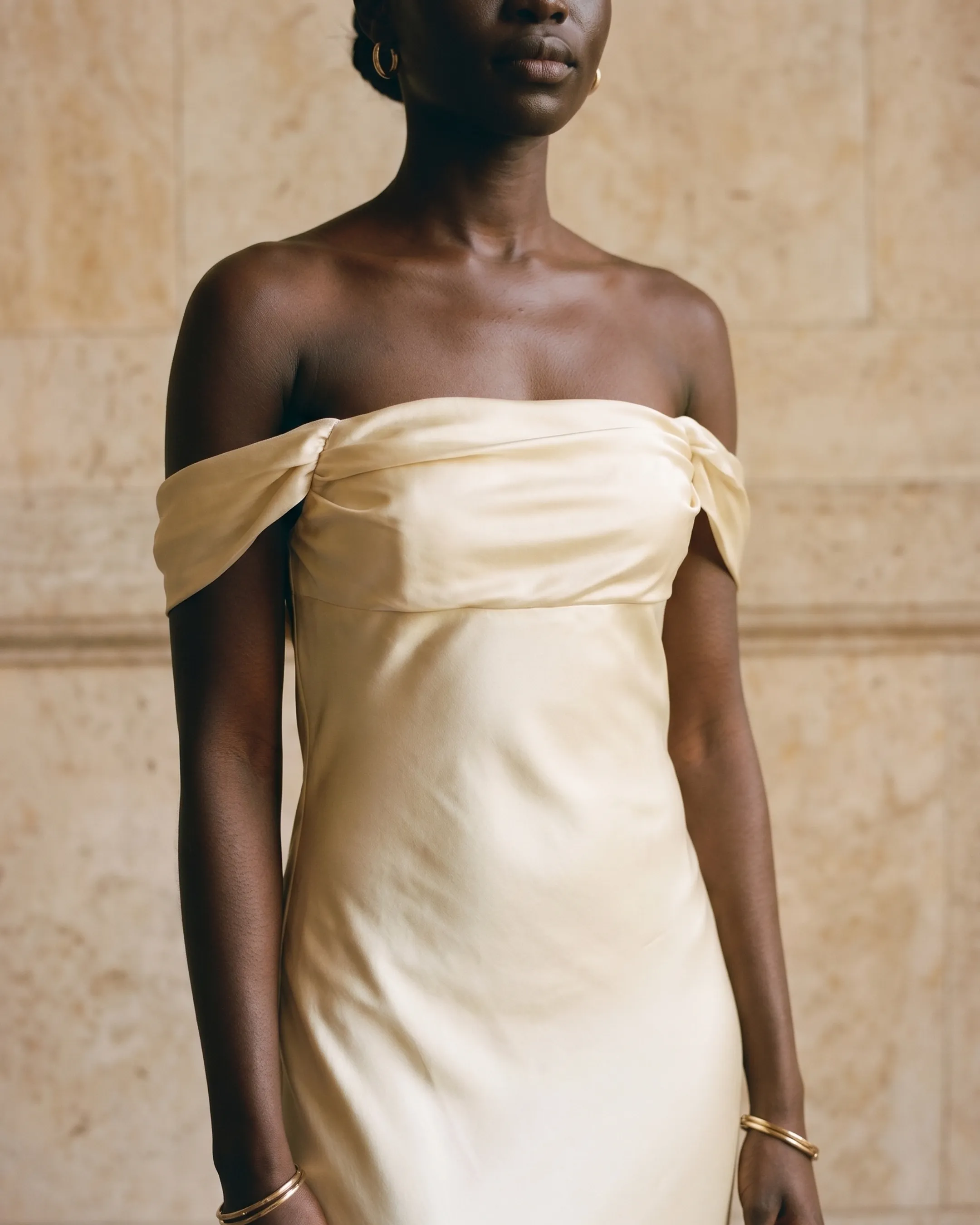 Off-the-shoulder draped satin bodice detail of the Hugo Caesar Tassi Flavia gown in champagne Avorio, lit by soft daylight against travertine marble