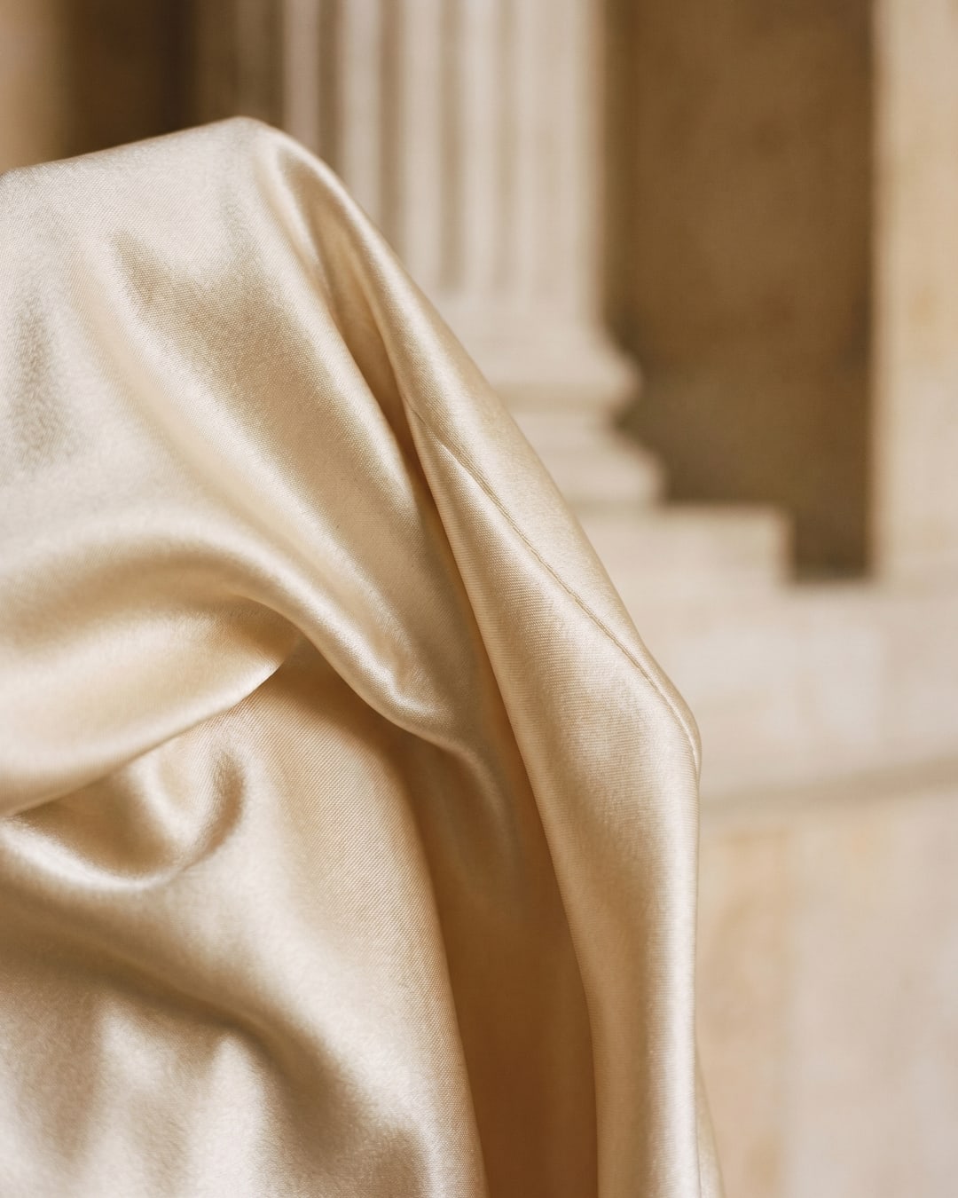 Macro detail of lustrous champagne liquid satin and fluid drape from the Hugo Caesar Tassi Flavia gown beside a fluted travertine column