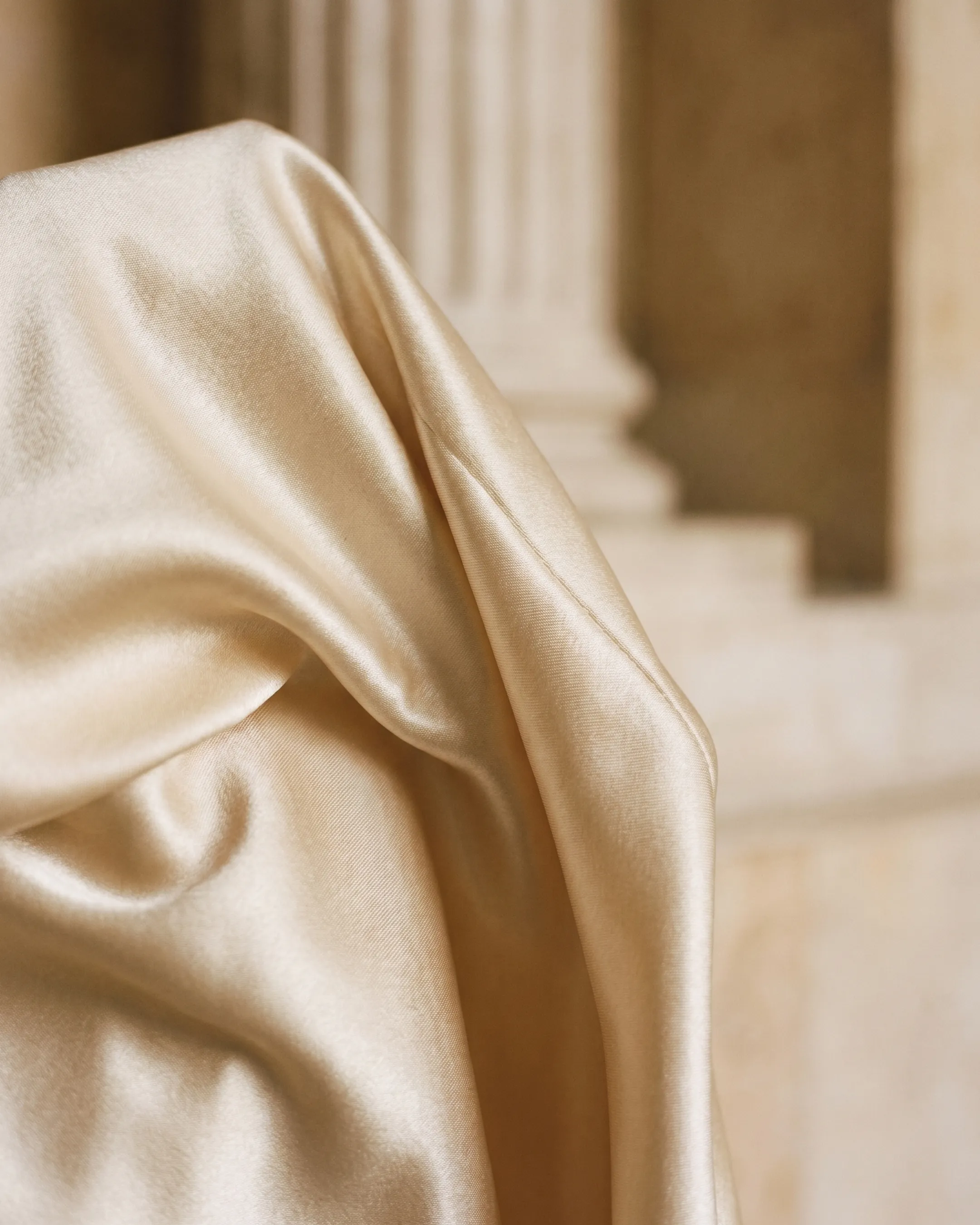 Macro detail of lustrous champagne liquid satin and fluid drape from the Hugo Caesar Tassi Flavia gown beside a fluted travertine column