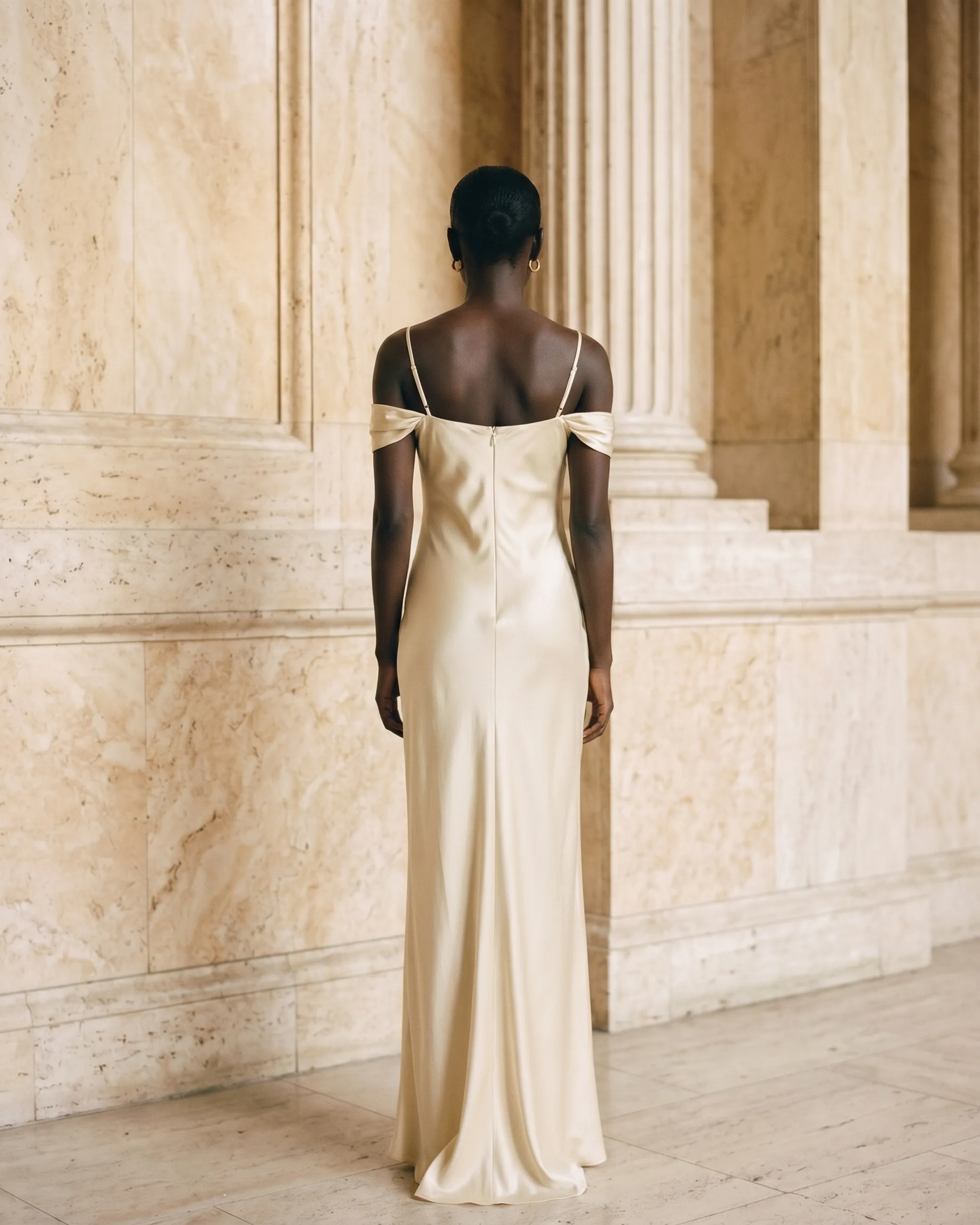 Full-length rear view of the Hugo Caesar Tassi Flavia strapless satin off-shoulder gown in champagne Avorio, seen from behind, showing the back of the dress, the bare off-shoulder back, concealed zip and liquid-satin column falling to the floor in a Roman travertine studio