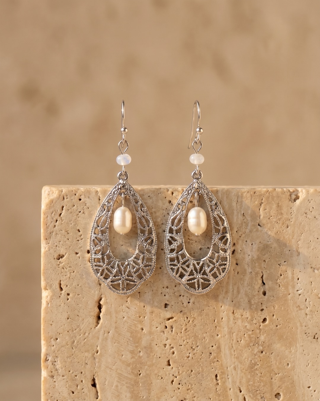 Hugo Caesar Tassi Fortuna pair of fine silver openwork teardrop drop earrings with a natural cream freshwater pearl, resting on a warm travertine pedestal