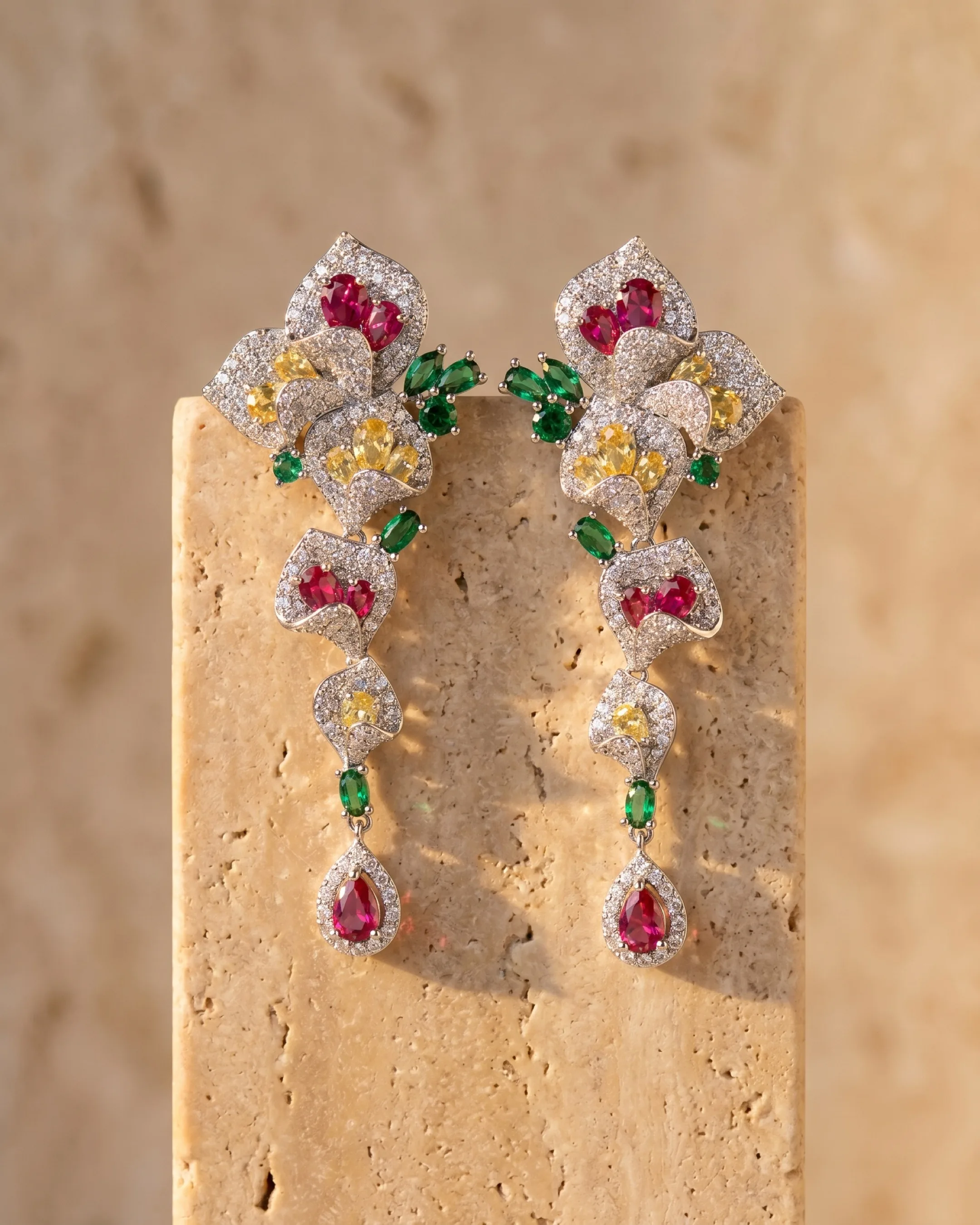 Hugo Caesar Tassi Gemma pair of long silver-set crystal cascade drop earrings with clear pave and coloured stones, resting against a warm travertine pedestal