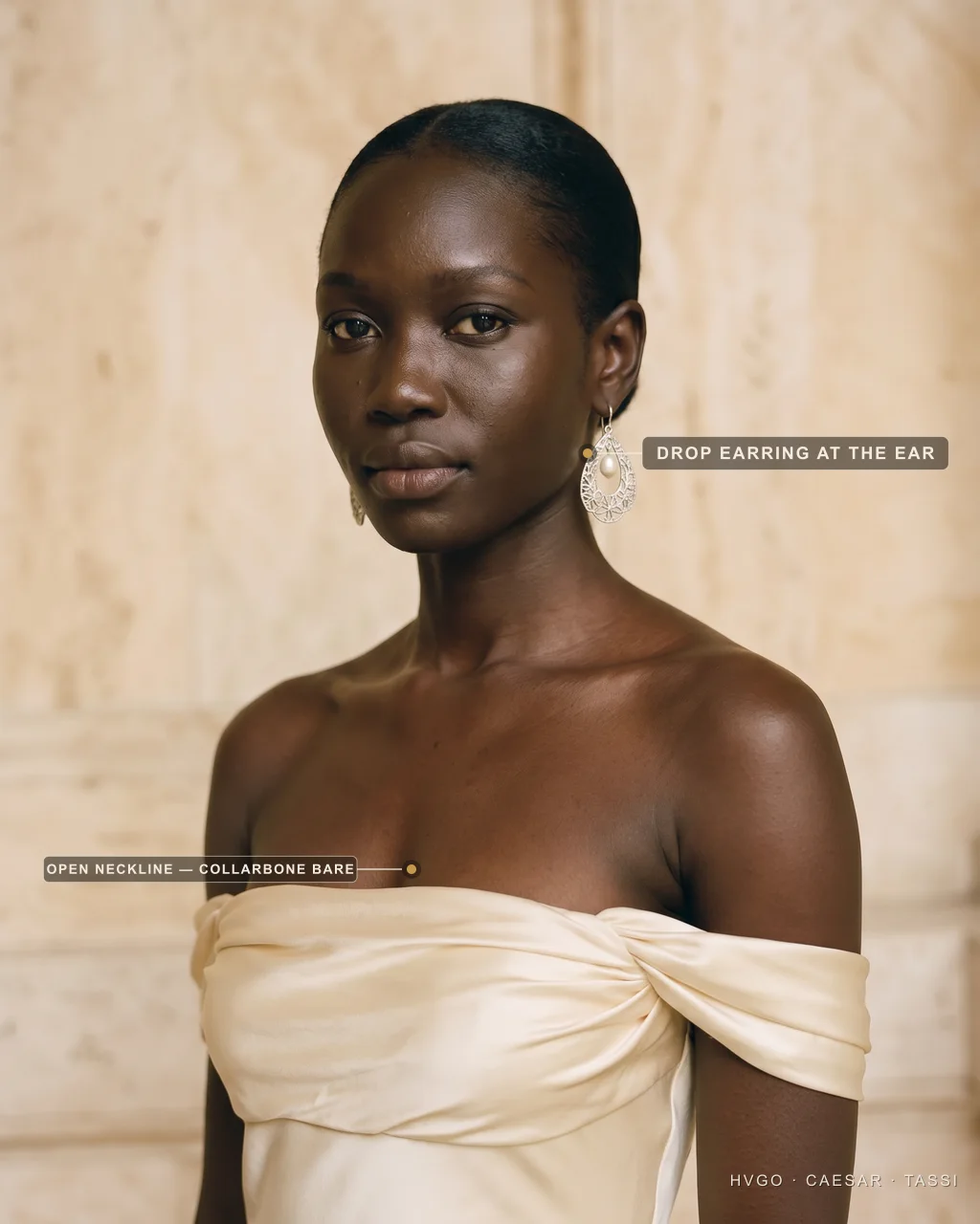 Close view of a woman in a Hugo Caesar Tassi champagne strapless off-the-shoulder satin gown wearing a silver filigree teardrop pearl drop earring, labelled to show the drop earring at the ear and the open neckline leaving the collarbone bare, in a Roman travertine studio
