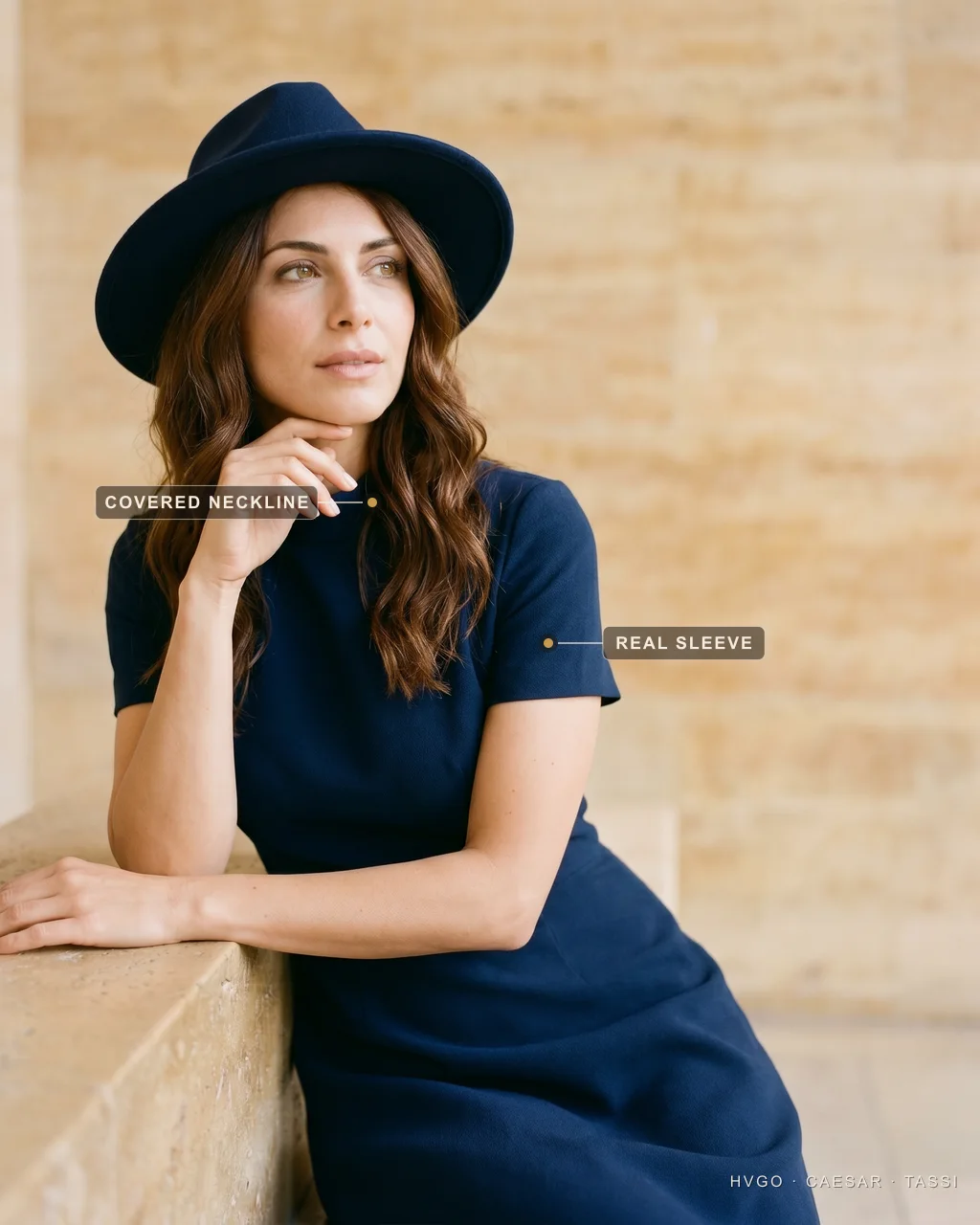 Royal Ascot short sleeve and covered neckline with a day hat — Hugo Caesar Tassi Sofia in a Hugo Caesar Tassi Blu Notte navy short-sleeve A-line crepe dress with a neat navy day hat, seated and leaning on a low travertine ledge at a three-quarter angle, labelled to show the covered high neckline and the real short sleeve, in a warm Roman travertine setting