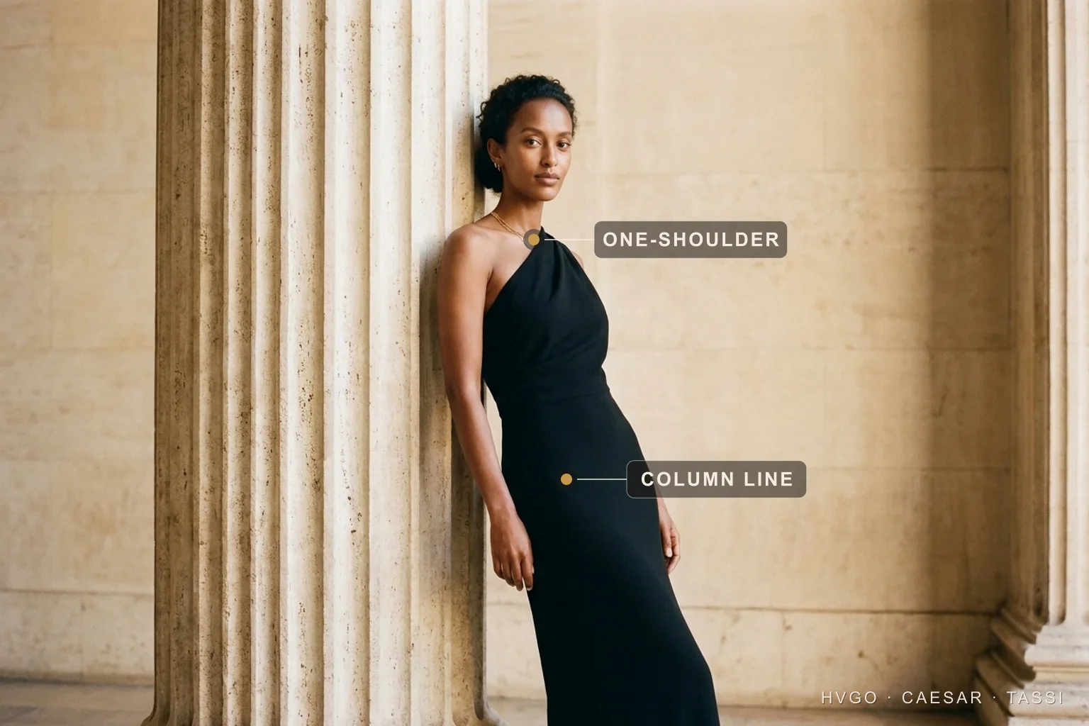 Black-tie column gown for women — the floor-length evening line — Hugo Caesar Tassi Aisha in a Hugo Caesar Tassi Basalt-black floor-length evening column gown, leaning against a fluted travertine column in a sunlit Roman colonnade, labelled to show the one-shoulder neckline at the strap and the unbroken column line falling down the gown body