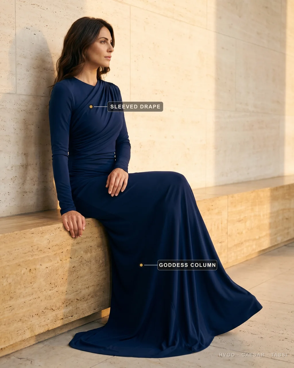 Black-tie draped goddess gown for women — sleeved, modest evening line — Hugo Caesar Tassi Sofia in a Hugo Caesar Tassi Cobalto-navy long-sleeved draped goddess column gown, seated and leaning on a low travertine ledge in profile in soft golden Roman light, labelled to show the sleeved Grecian drape at the shoulder and the goddess column pooling at the hem