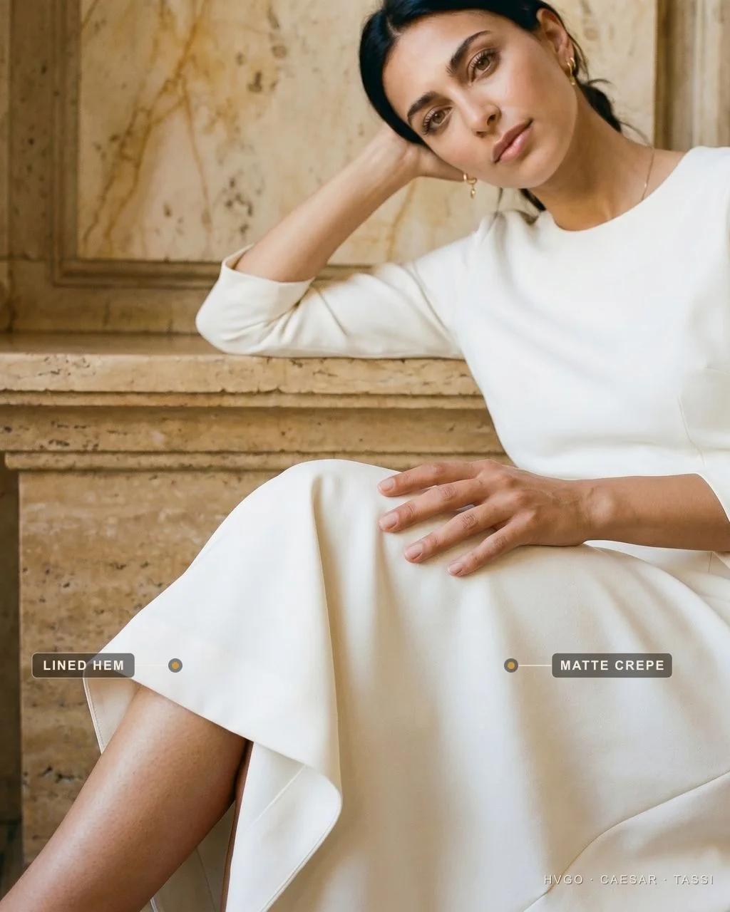 Matte crepe and a fully-lined hem — Hugo Caesar Tassi Close detail of a seated model's ivory dress, with baked labels marking the structured matte-crepe fabric across the lap and the clean fully-lined hem fold over the knee.