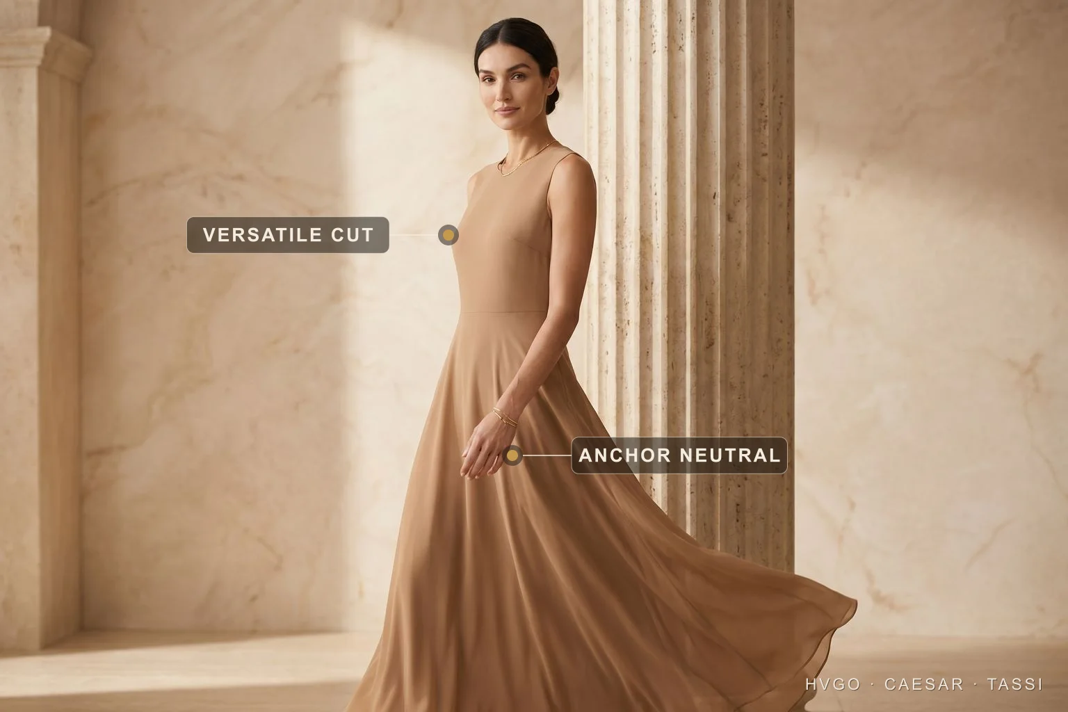 A neutral anchor dress that recombines — Hugo Caesar Tassi A model in a camel-stone A-line chiffon maxi dress walking beside a travertine column, with baked labels marking the versatile cut at the bodice waist and the anchor neutral colour on the skirt.