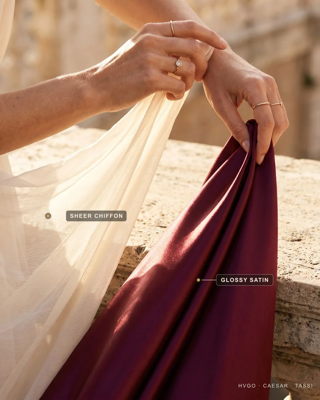 Sheer chiffon vs glossy satin — fabric surfaces compared | Hugo Caesar Tassi Close-up of two hands draping two fabrics over a travertine ledge: translucent ivory chiffon on the left and glossy wine satin on the right, with baked labels reading Sheer chiffon and Glossy satin.