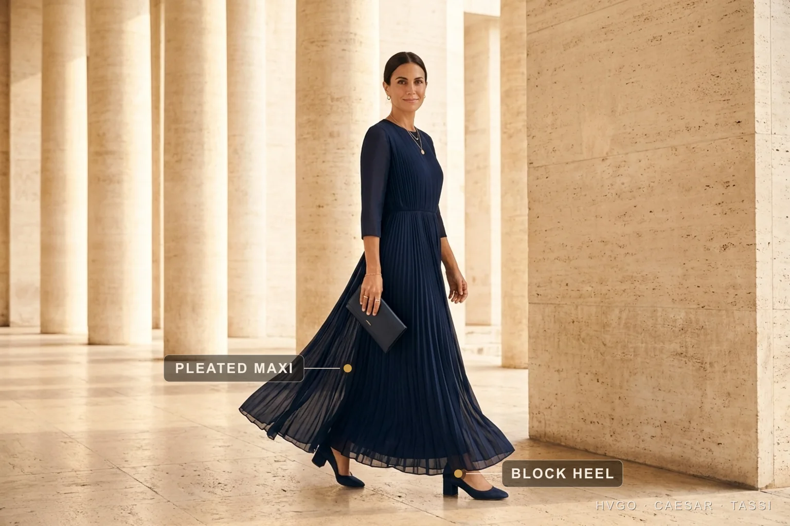 Godmother Christening Outfit: a Navy Pleated Chiffon Maxi — Hugo Caesar Tassi Marisol, a dark-haired house model, walking mid-stride through a Roman travertine colonnade in a Hugo Caesar Tassi Cobalto deep-navy French-pleated chiffon maxi with a slim clutch, labelled to show the fluttering pleated maxi skirt and the low block heel built for standing at the font