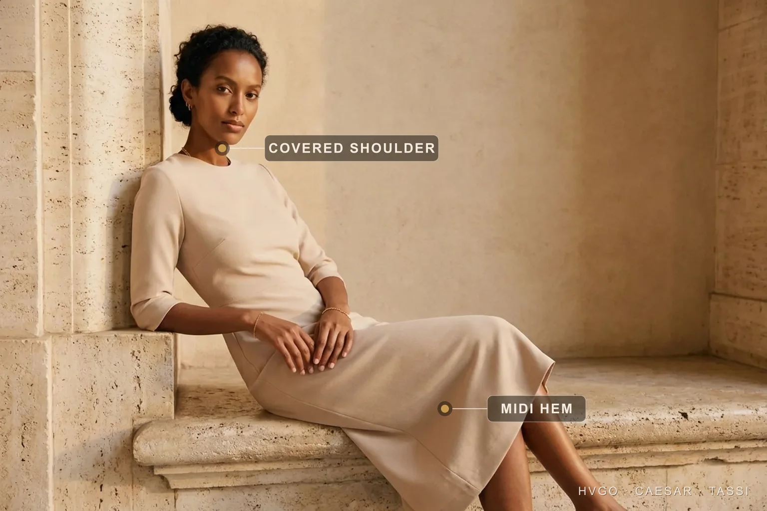 What to Wear to a Christening: a Modest Stone-Crepe Midi — Hugo Caesar Tassi Aisha, a house model with natural dark curls, seated and leaning three-quarters on a sunlit Roman travertine ledge in a Hugo Caesar Tassi soft stone-beige matte-crepe christening midi, labelled to show the covered shoulder and three-quarter sleeve and the knee-clearing midi hem