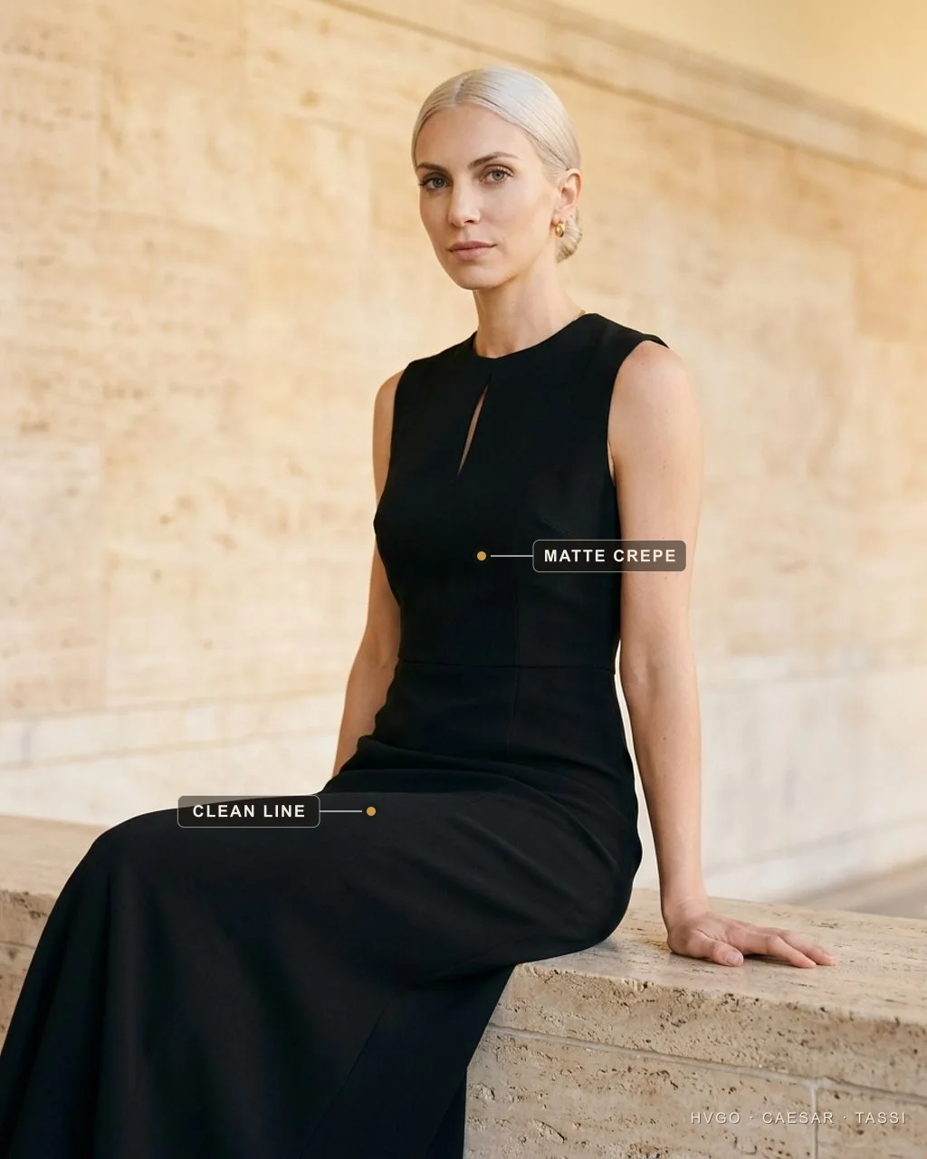 A tailored crepe cocktail sheath for women — Hugo Caesar Tassi Valentina seated and leaning on a low travertine ledge in a Hugo Caesar Tassi black tailored matte heavy-crepe cocktail sheath with a single collarbone cutout, labelled to show the matte crepe and its clean tailored line, in a warm Roman travertine setting
