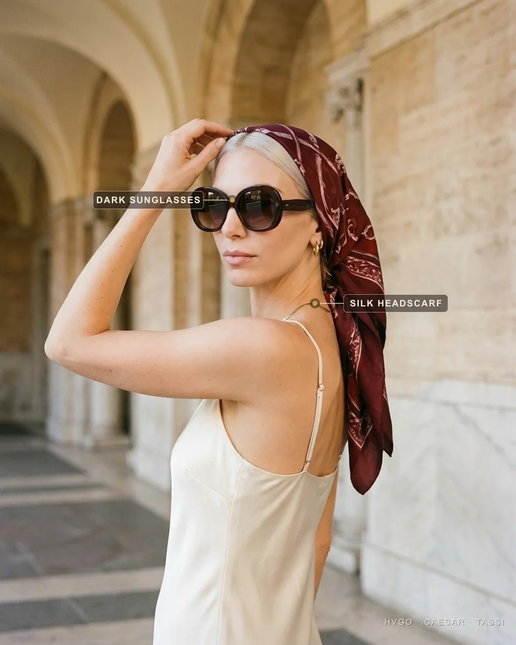 Valentina, a platinum-blonde Hugo Caesar Tassi house model in an ivory satin slip, glancing back over her shoulder with one hand raised to an oxblood-and-wine printed silk square tied as a headscarf and oversized dark sunglasses, labelled to show the dark sunglasses and the silk headscarf, in a sunlit Roman travertine arcade