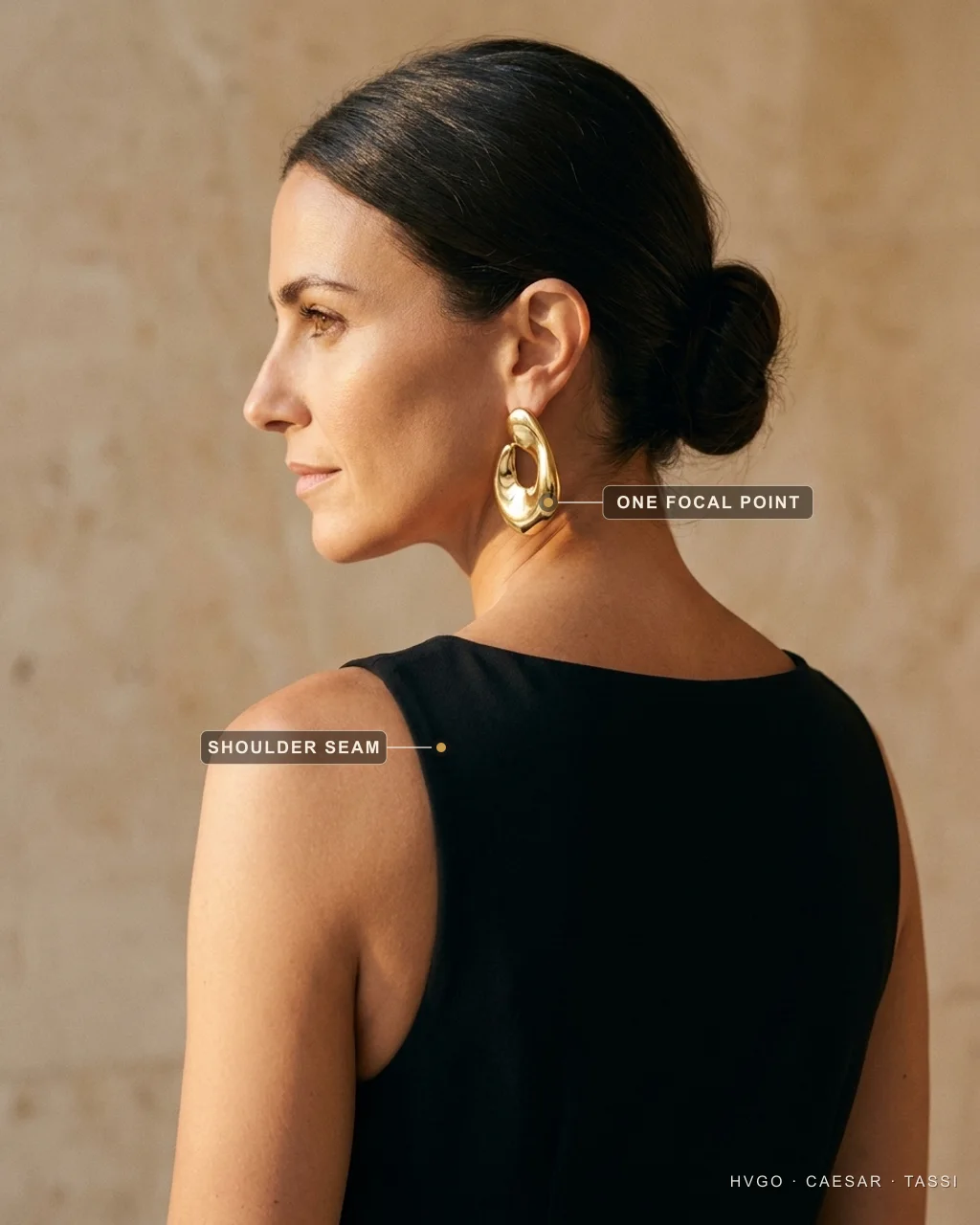 How to dress elegantly: shoulder-seam fit and one focal point — Hugo Caesar Tassi Marisol, a Hugo Caesar Tassi house model, in three-quarter-back profile in a Basalt-black sleeveless crepe dress wearing one oversized polished-gold sculptural statement earring, labelled to show the earring as the single focal point and the shoulder seam set at the shoulder's edge, in a warm Roman travertine setting