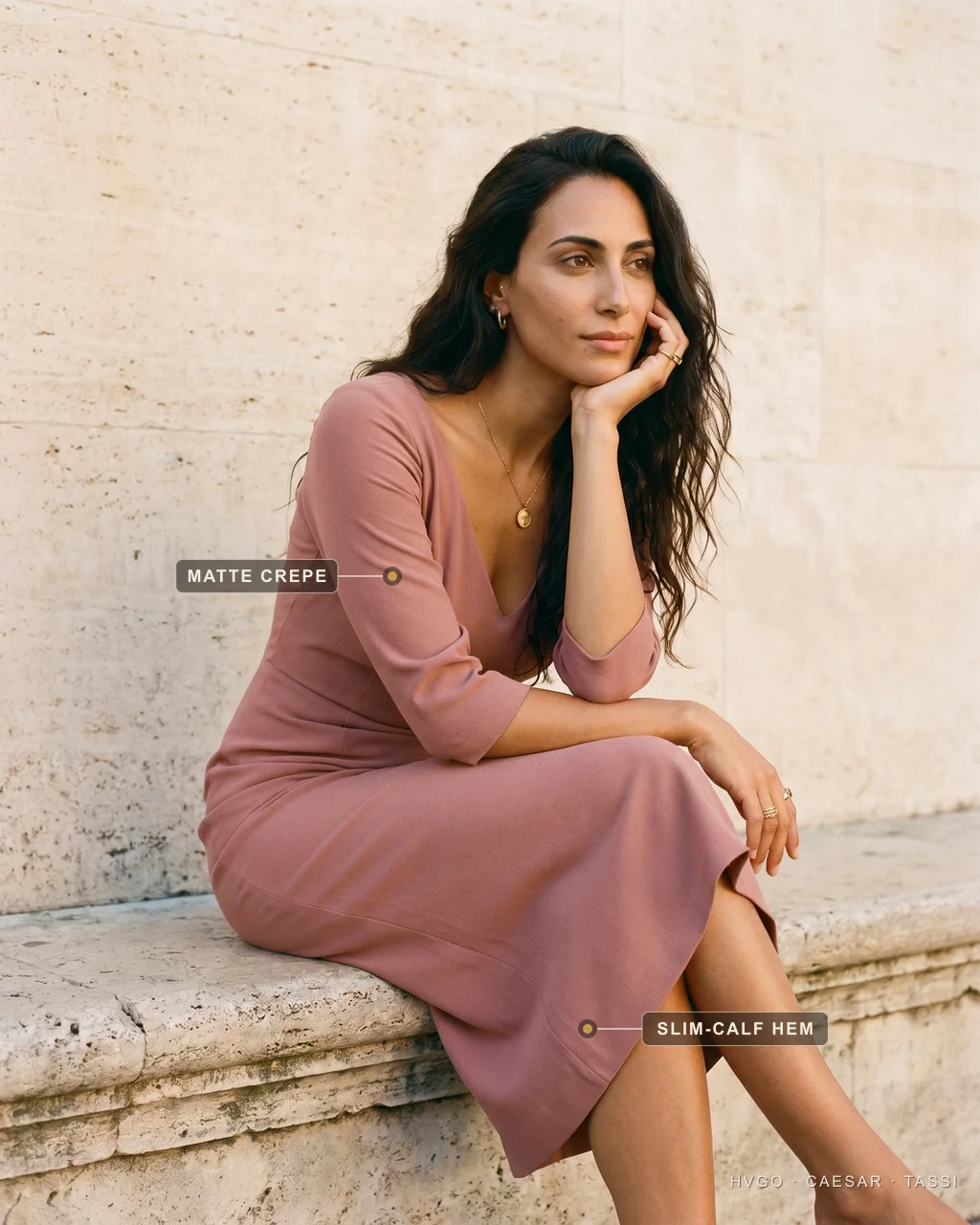 How to dress elegantly: matte crepe and a slim-calf hem — Hugo Caesar Tassi Chiara, a Hugo Caesar Tassi house model, seated on a travertine ledge with a hand to her cheek in a dusty-rose Cornelia matte-crepe dress, labelled to show the matte crepe and the slim-calf hem, in warm Roman daylight