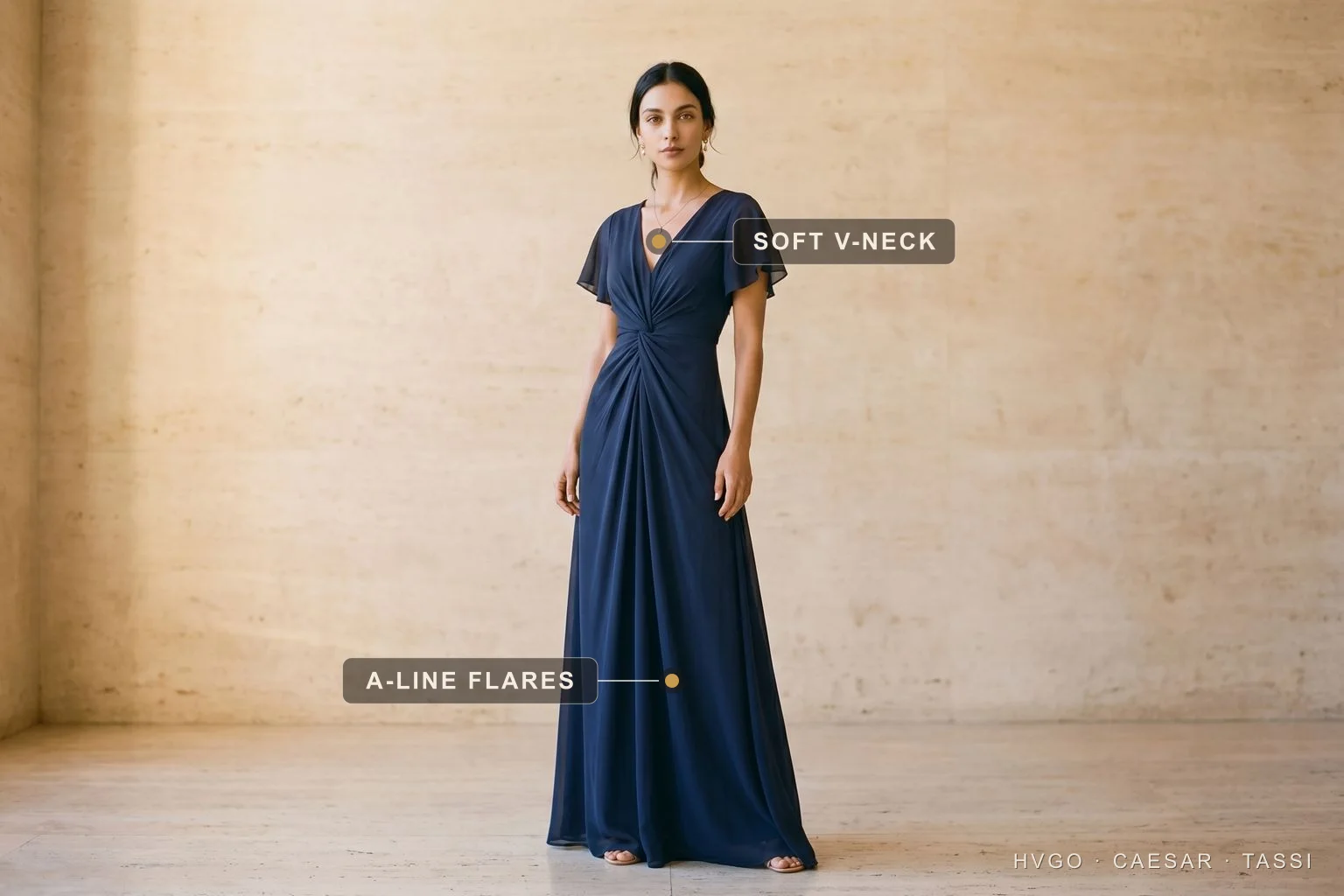 A woman in a Hugo Caesar Tassi cobalt-navy chiffon A-line gown with a soft V-neckline and flutter sleeves in a Roman travertine studio, labelled to show the soft V-neckline and the A-line skirt flaring to the hem