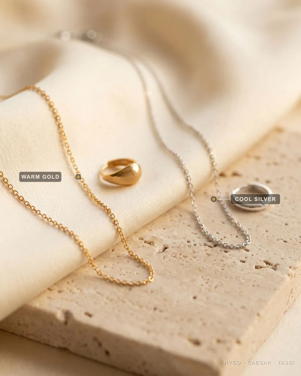 Flat-lay of fine jewelry showing warm yellow-gold chain and ring on ivory silk beside a cool sterling-silver chain and hoop on travertine, each metal labelled