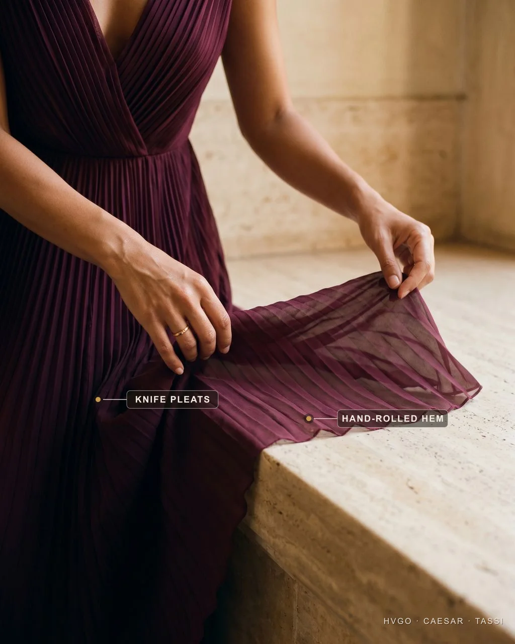 No-face detail of model Chiara's hands laying the knife-pleated chiffon skirt of the Cassia V-Neck Pleated Chiffon Gown in porpora wine over a travertine ledge, the translucent chiffon catching golden light; labels mark the Knife pleats and the Hand-rolled hem.