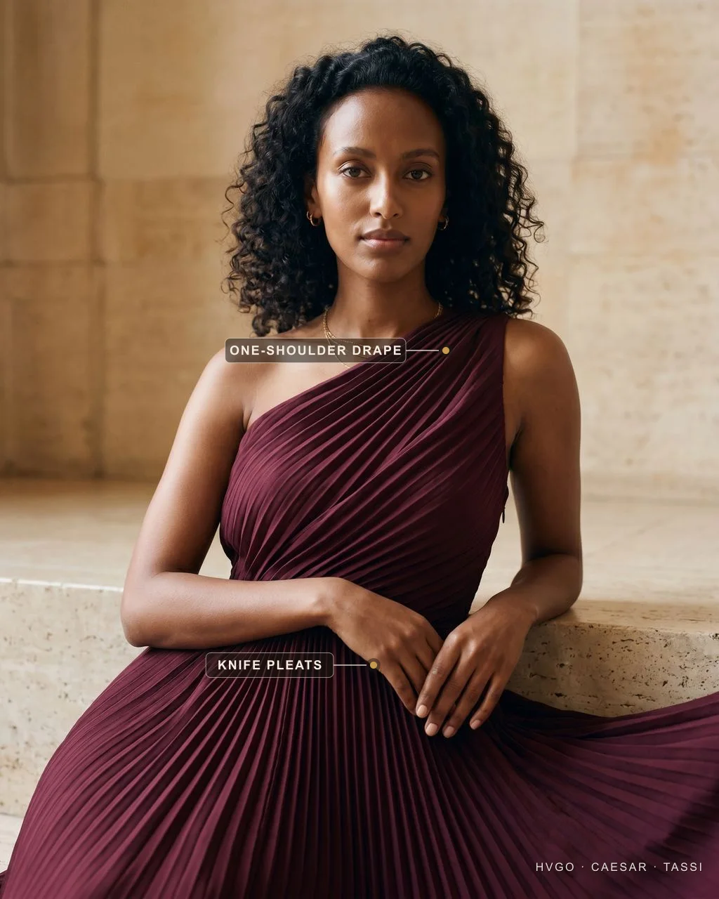 A second model seated and leaning on a travertine ledge in the Hugo Caesar Tassi Tullia pleated column evening gown in deep Porpora wine crepe; gilt labels mark the one-shoulder drape and the knife-pleated skirt.