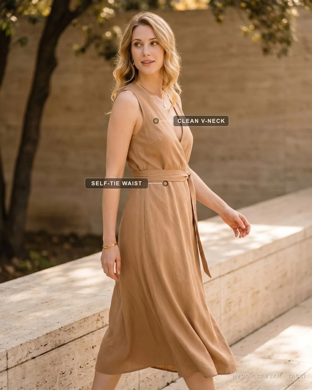 Italian summer style, a Cuoio tencel-linen self-tie wrap | Hugo Caesar Tassi Northern-Italian model walking mid-stride in a camel-stone Cuoio Lavinia sleeveless V-neck self-tie wrap dress in tencel-linen by a travertine garden ledge; gilt labels mark the clean V-neckline and the self-tie waist sash.