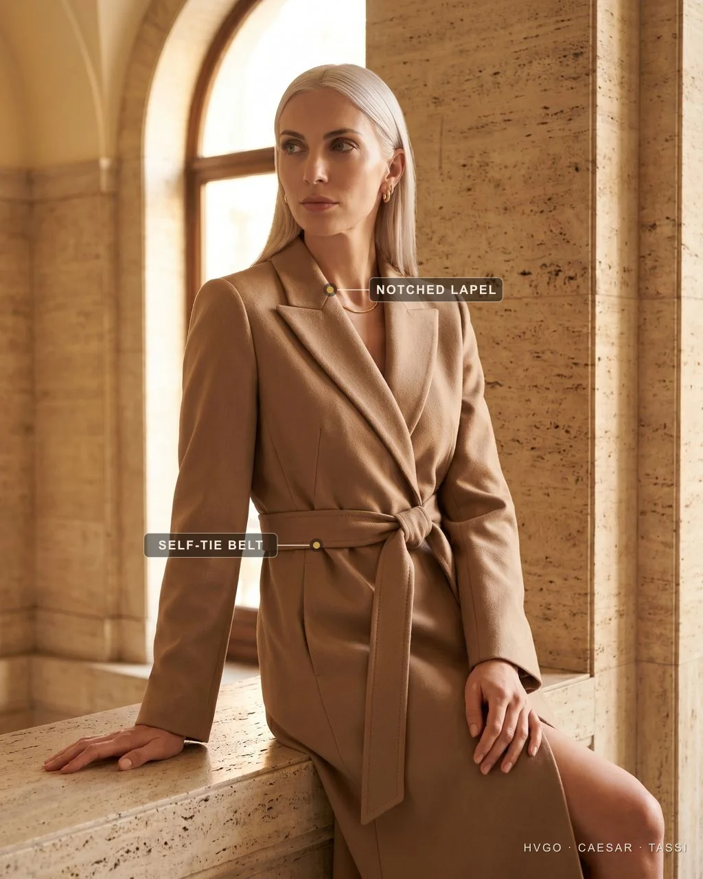 Platinum-blonde woman seated on a travertine ledge in the Hugo Caesar Tassi Drusilla belted tailored coat-dress in camel-stone, with labels marking the notched lapel and the self-tie belt.