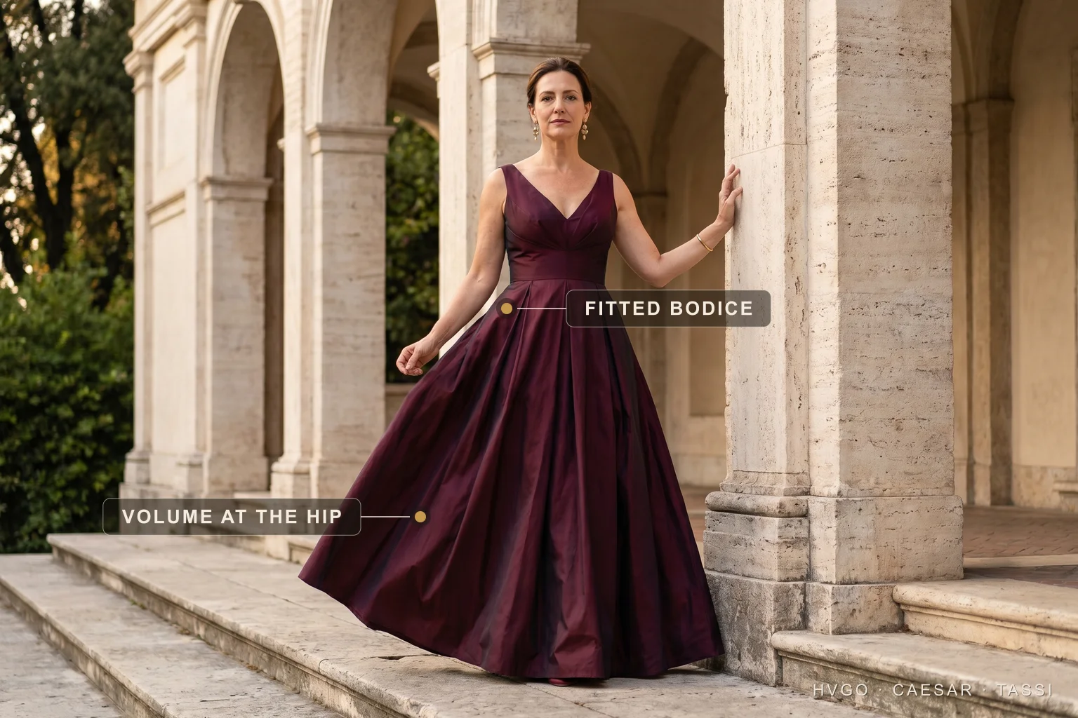 Fit-and-flare gown for an inverted-triangle figure — Hugo Caesar Tassi A woman with an inverted-triangle figure in a Hugo Caesar Tassi wine fit-and-flare gown in a Roman loggia, labelled to show the fitted V-neck bodice and the pleated skirt building hip volume
