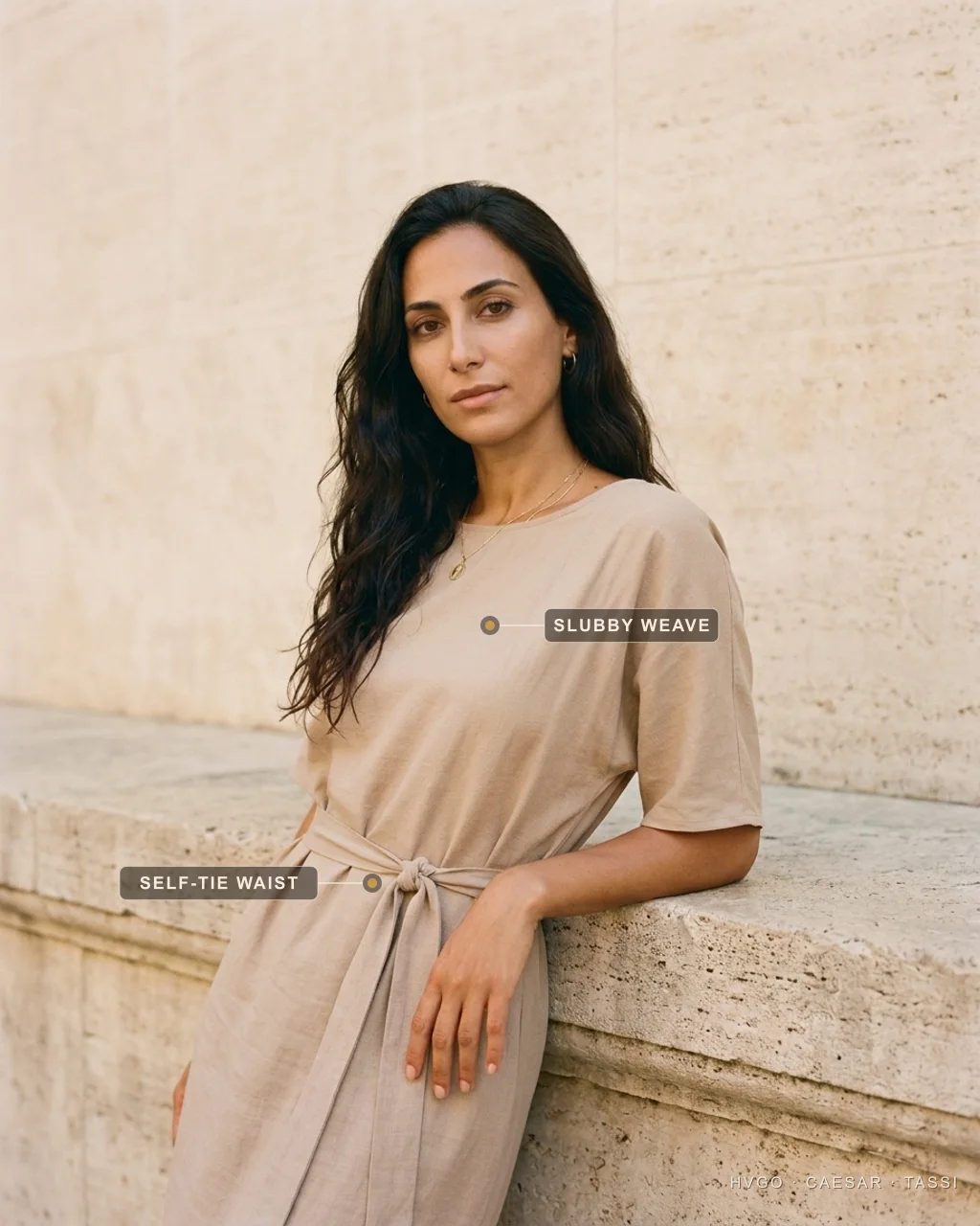 Close portrait of a dark-haired woman in a Hugo Caesar Tassi Travertine stone-beige Tencel-linen day dress, leaning a forearm on a Roman travertine ledge in a three-quarter turn, labelled to show the matte slubby Tencel-linen weave at the chest and the soft self-tie knotted at the waist