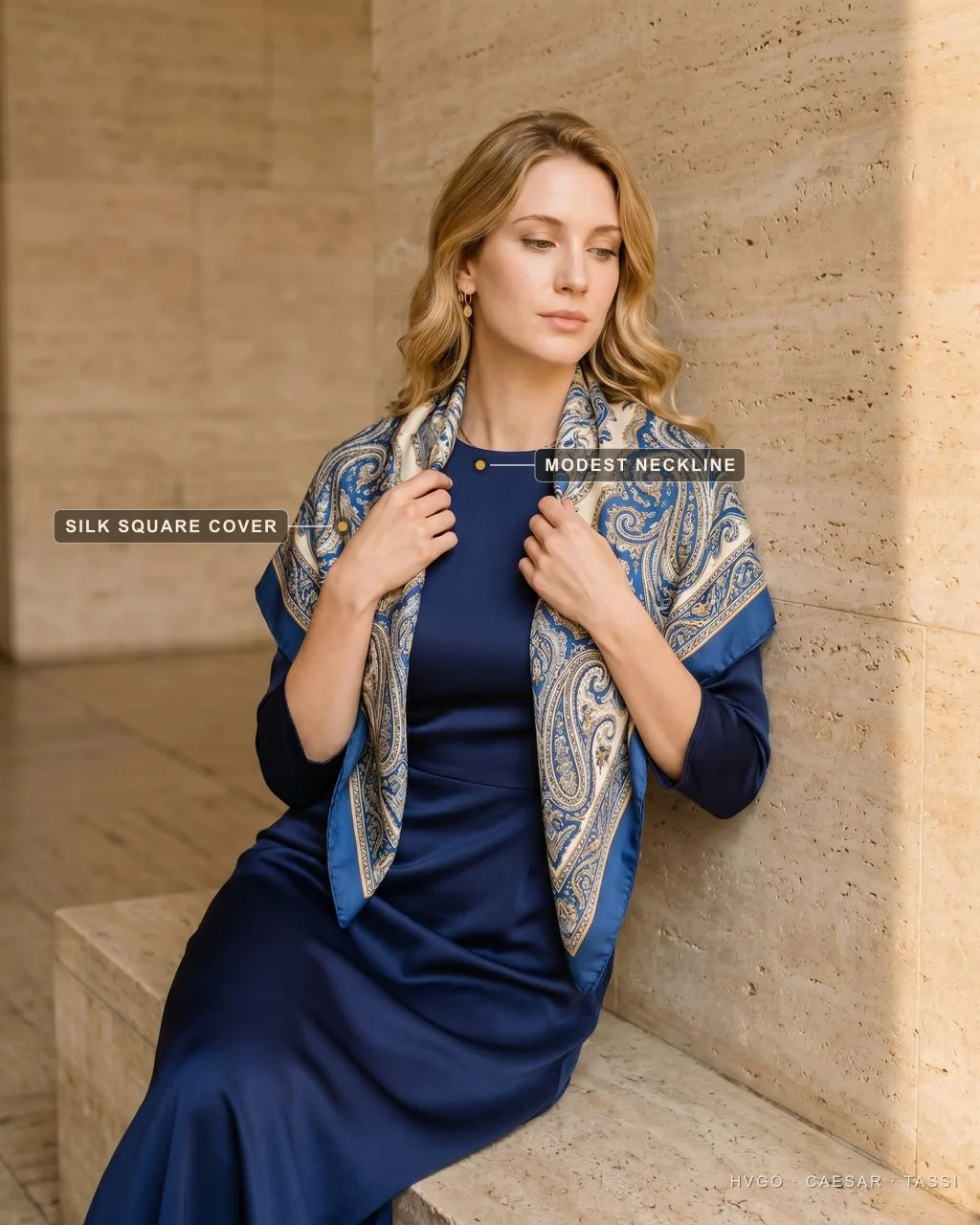 Silk-square church cover-up over a navy dress — Hugo Caesar Tassi Italian wedding guest Bianca seated and leaning on a travertine ledge in a Cobalto navy satin-crepe dress, a large patterned silk square draped over both shoulders as a church cover-up, one hand adjusting the scarf, labelled to show the silk-square cover and the modest neckline in a warm Roman setting — the elegant chiesa solution