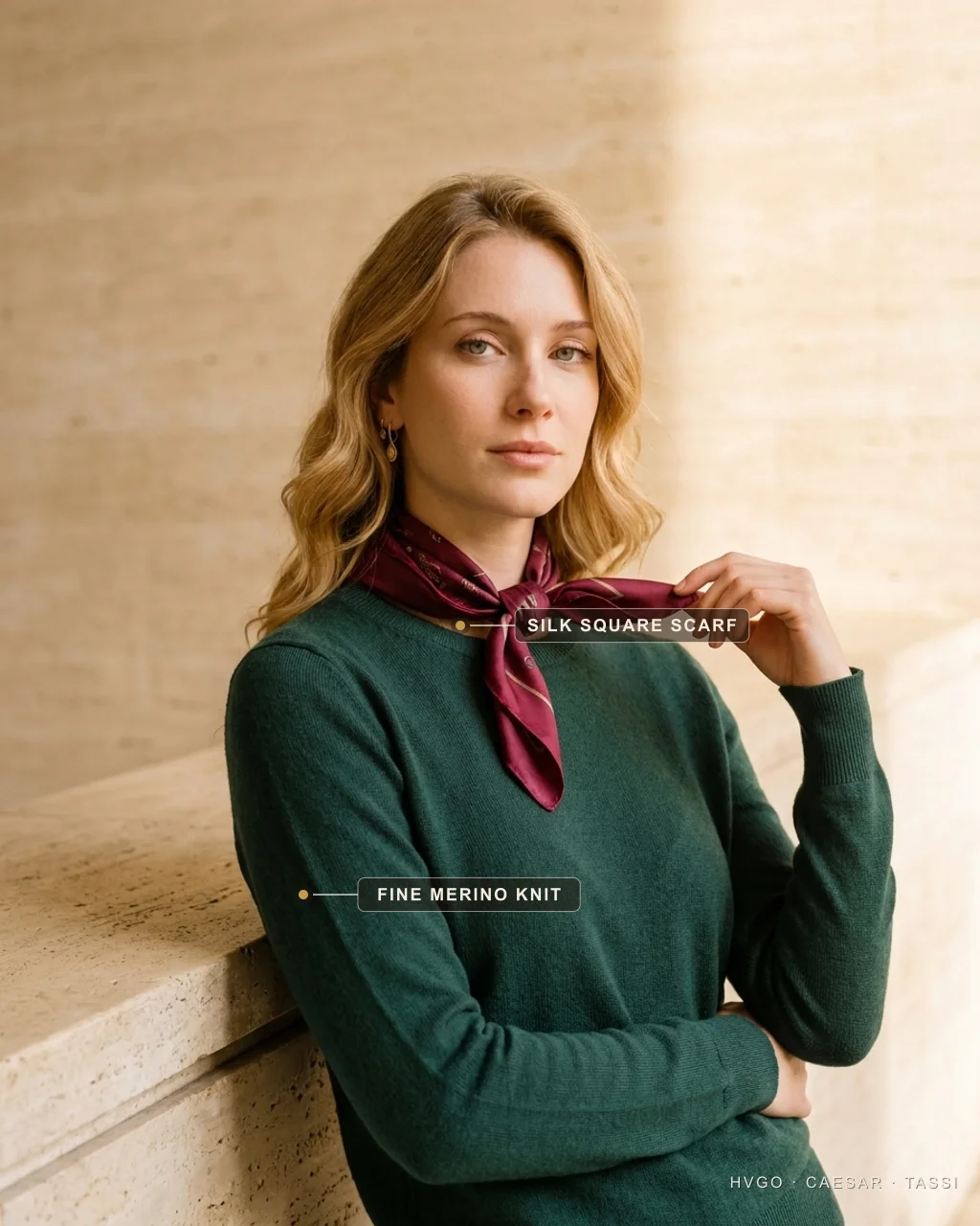 Bianca, a golden-blonde Italian woman, in three-quarter turn leaning on a warm travertine ledge, wearing a Hugo Caesar Tassi fine-gauge Verde-Antico green merino knit with a Porpora wine silk-twill square scarf knotted at the throat as the one rich accent, one hand lifting the scarf edge, labelled to show the silk square scarf and the fine merino knit, in a warm Roman travertine setting