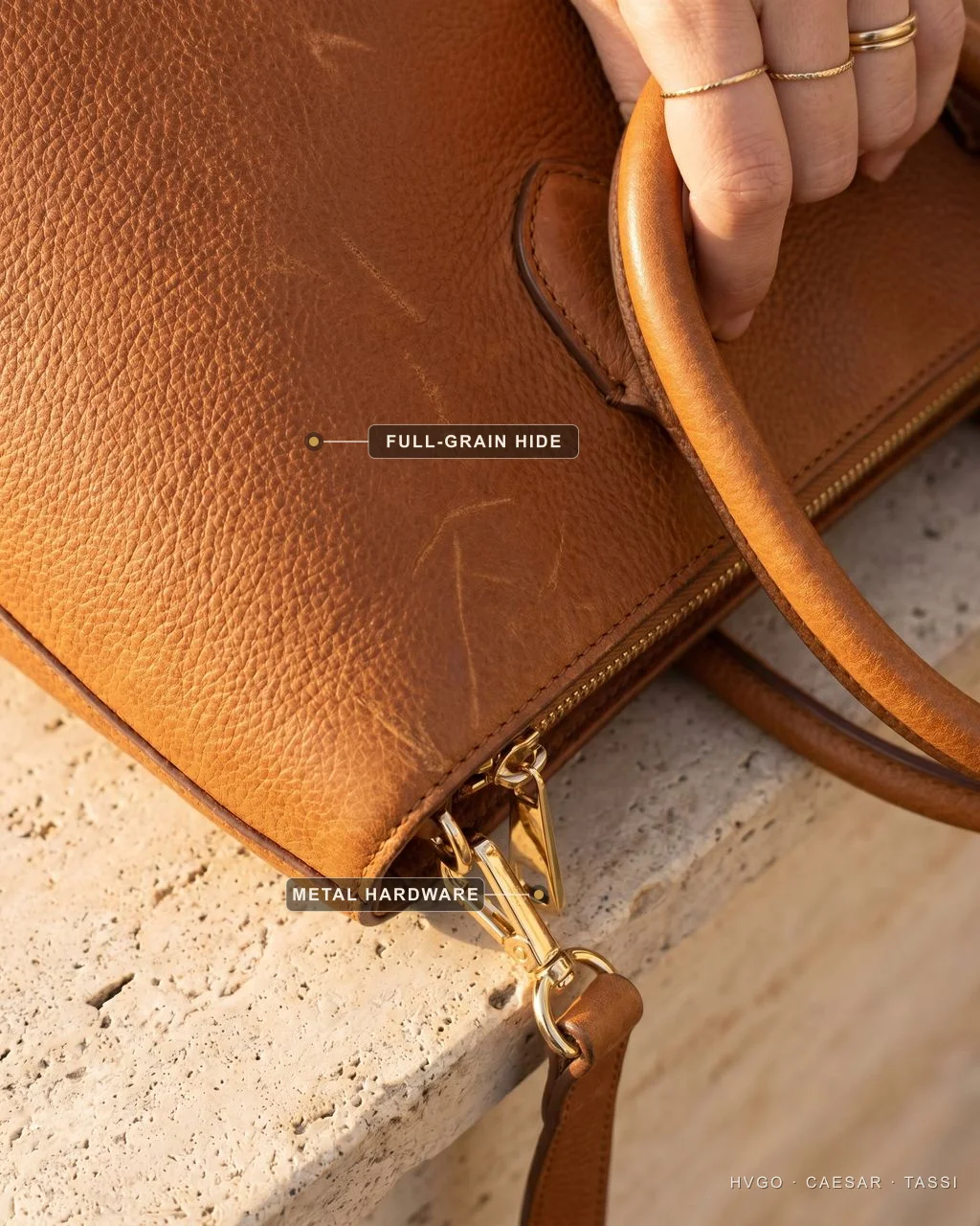 Macro close-up of a tan leather tote held by a hand, with labels marking the natural full-grain hide texture and the polished gold-tone metal hardware at the base.