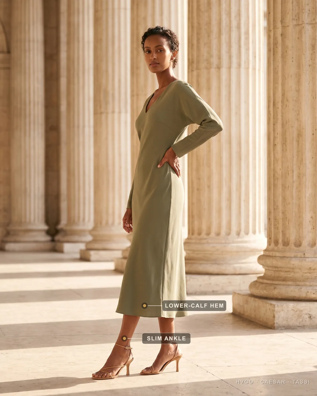 Midi Dress Length: The Hem Breaking at the Lower Calf — Hugo Caesar Tassi Aisha in a three-quarter contrapposto turn with one hand on her hip, wearing an olive sage-green crepe midi against warm Roman travertine columns, the baked markers showing the lower-calf hem and her slim ankle in a heeled sandal.