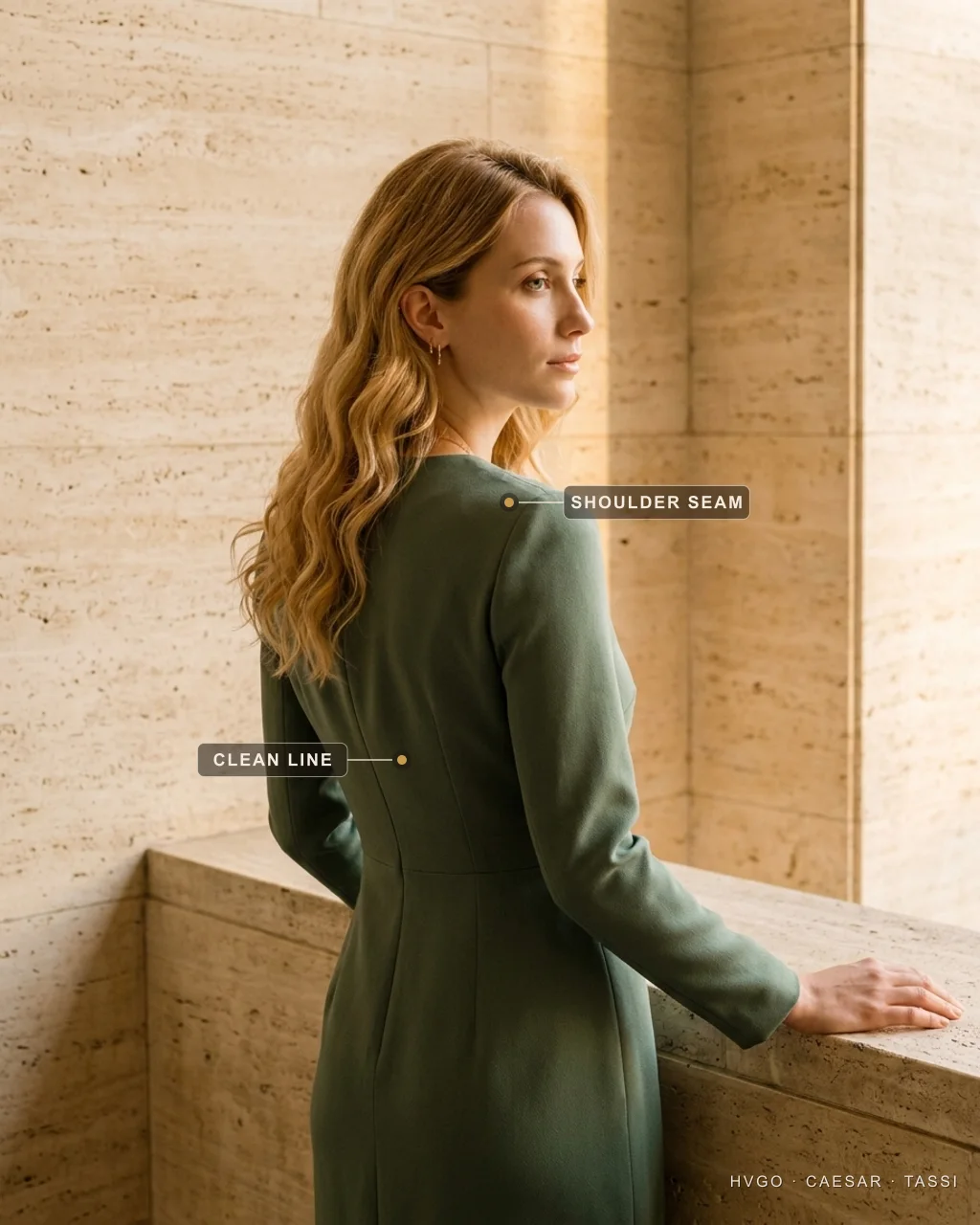How to look expensive: a clean shoulder line in a green crepe dress — Hugo Caesar Tassi Bianca, a golden-blonde Hugo Caesar Tassi house model, in a tailored Verde Antico green structured-crepe dress seen in three-quarter-back profile with one hand on a travertine ledge, labelled to show the set-in shoulder seam sitting at the bony shoulder point and the clean vertical back seam, in a sunlit Roman travertine corner