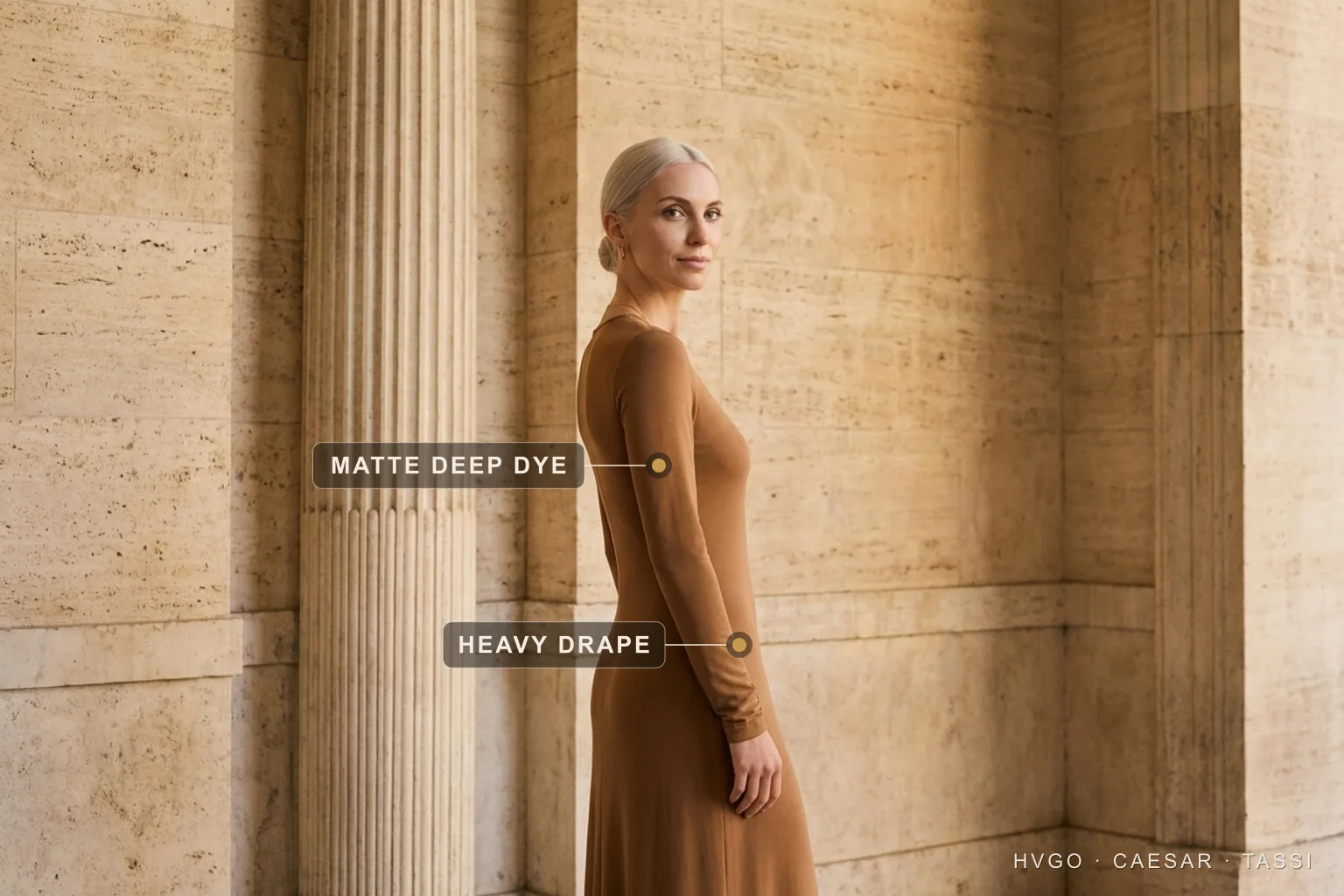 How to look expensive: drape and depth of dye in a tan column gown — Hugo Caesar Tassi Valentina, a platinum-blonde Hugo Caesar Tassi house model, in a deep Cuoio tan matte-modal column gown turned in three-quarter contrapposto beside a fluted travertine column, labelled to show the matte deep dye on the sleeve and the heavy drape falling in one fluid line, in a warm Roman travertine colonnade