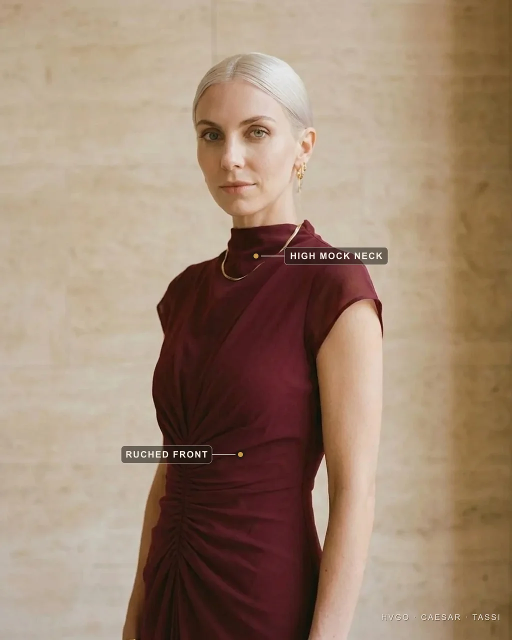 Dress necklines explained — the high mock neck — Hugo Caesar Tassi Close view of a woman in a Hugo Caesar Tassi wine mock-neck pleated gown, labelled to show the high columnar mock neck rising to a clean band at the throat and the ruched centre front, in a Roman travertine studio
