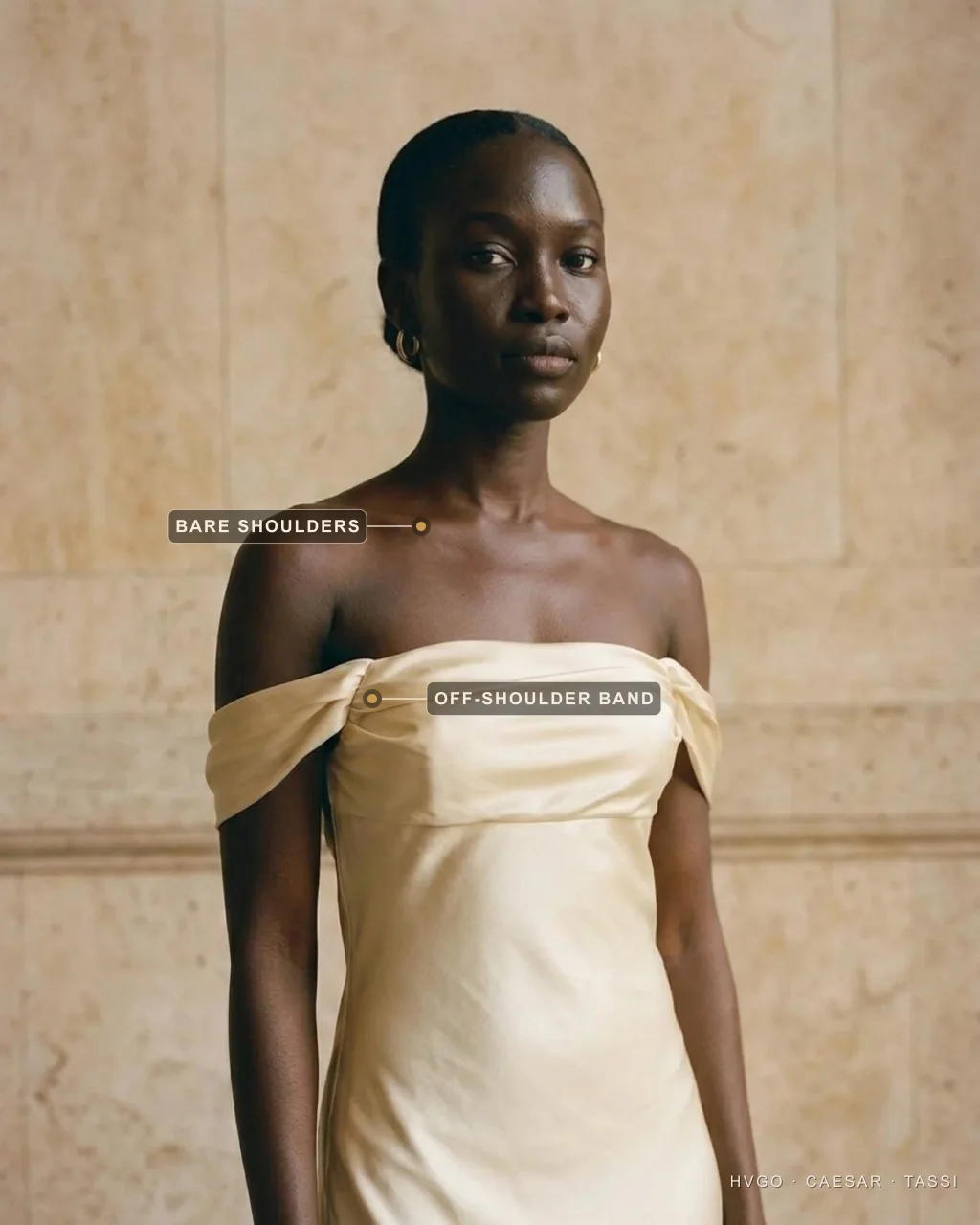 Dress necklines explained — the off-the-shoulder Bardot neckline — Hugo Caesar Tassi Close view of a woman in a Hugo Caesar Tassi champagne strapless off-the-shoulder satin gown, labelled to show the Bardot band sitting off the shoulders and the bare shoulders and collarbone, in a Roman travertine studio