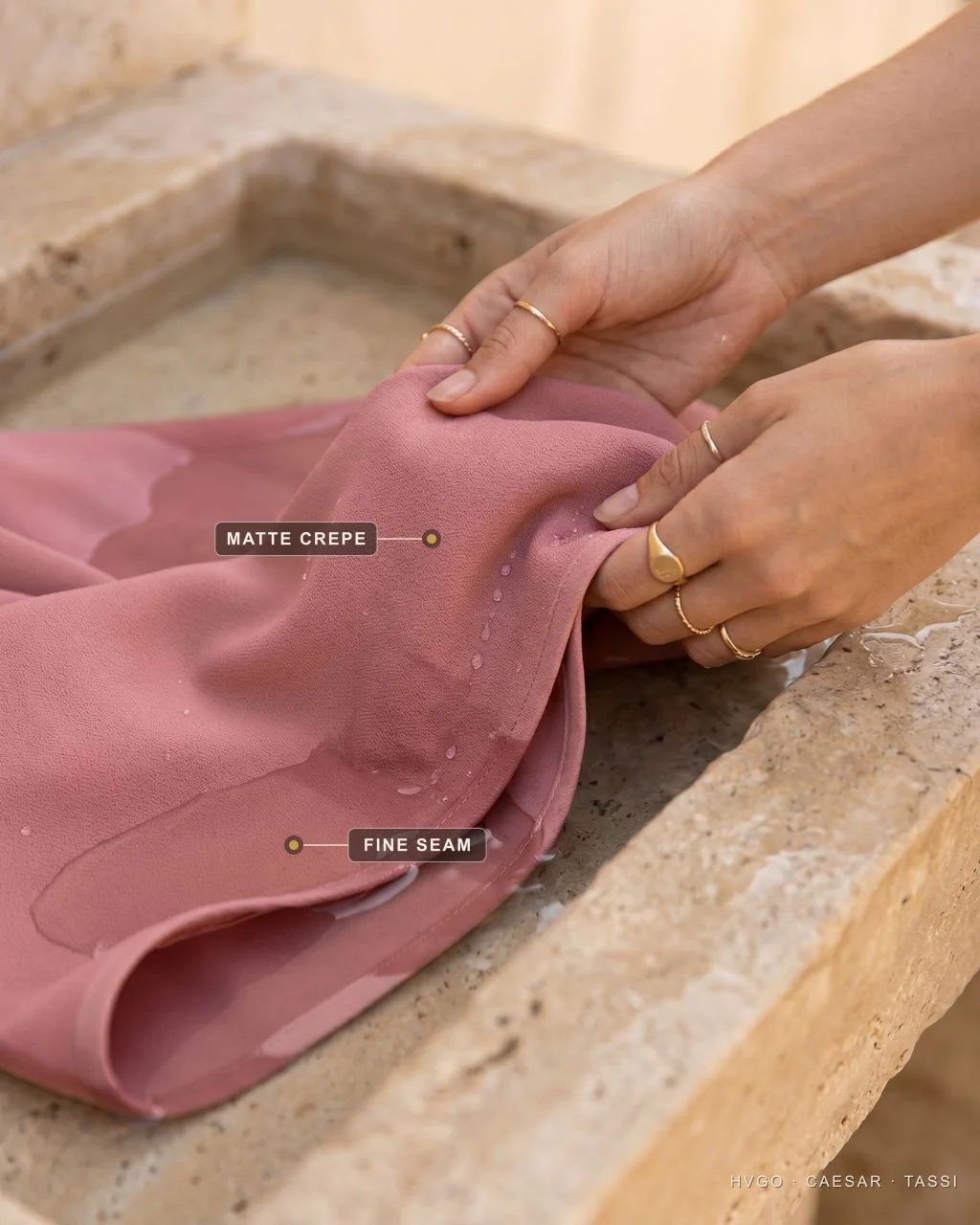 Hand-washing a crepe occasion dress — Hugo Caesar Tassi Close-up of hands gently swishing a Cipria-rose crepe dress in a shallow travertine basin of cool water, with markers on the matte crepe surface and the fine seam at the folded hem edge.