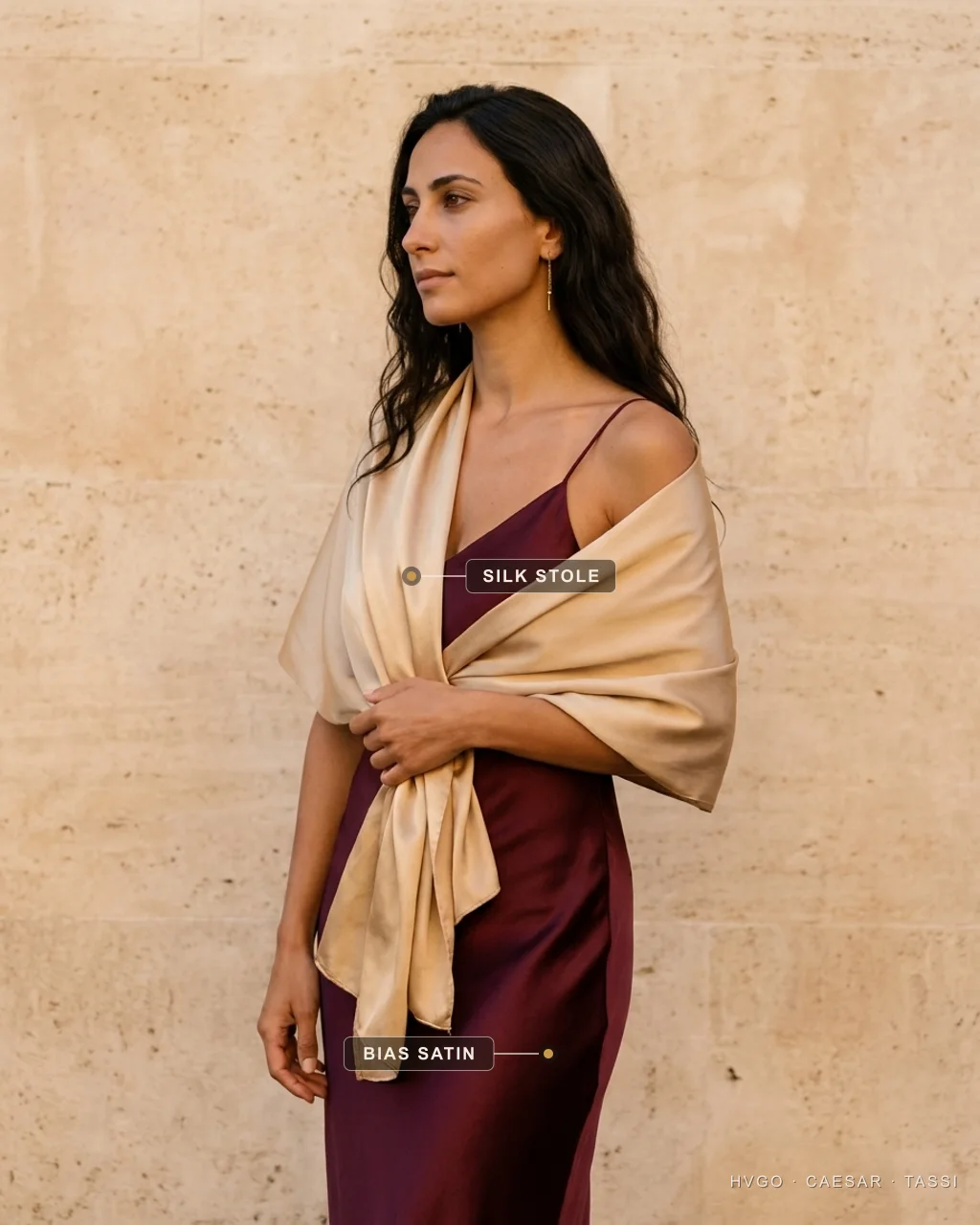 Chiara, an HCT house model with dark long waves, shown in three-quarter profile in a champagne-gold silk stole wrapped over the shoulders and arms above a deep Porpora-wine bias-cut satin gown, labelled to show the silk stole and the bias-satin gown beneath, in a warm Roman travertine setting